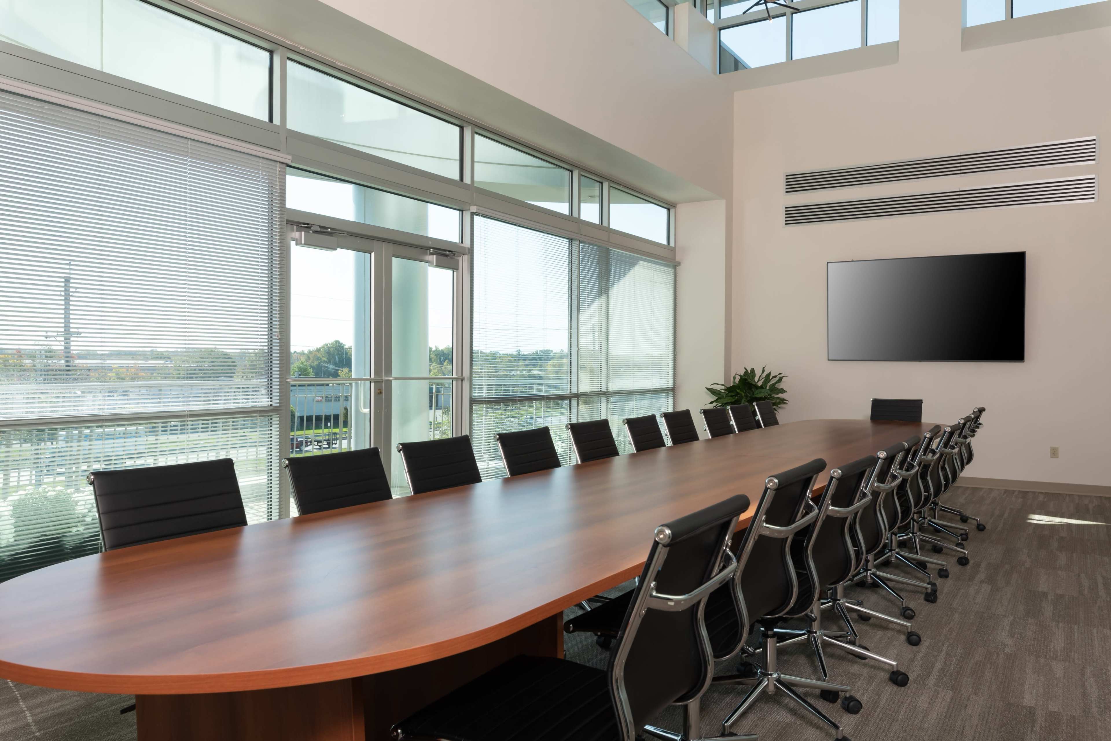 A large conference room features a wooden table surrounded by black chairs, with floor-to-ceiling windows letting in natural light.