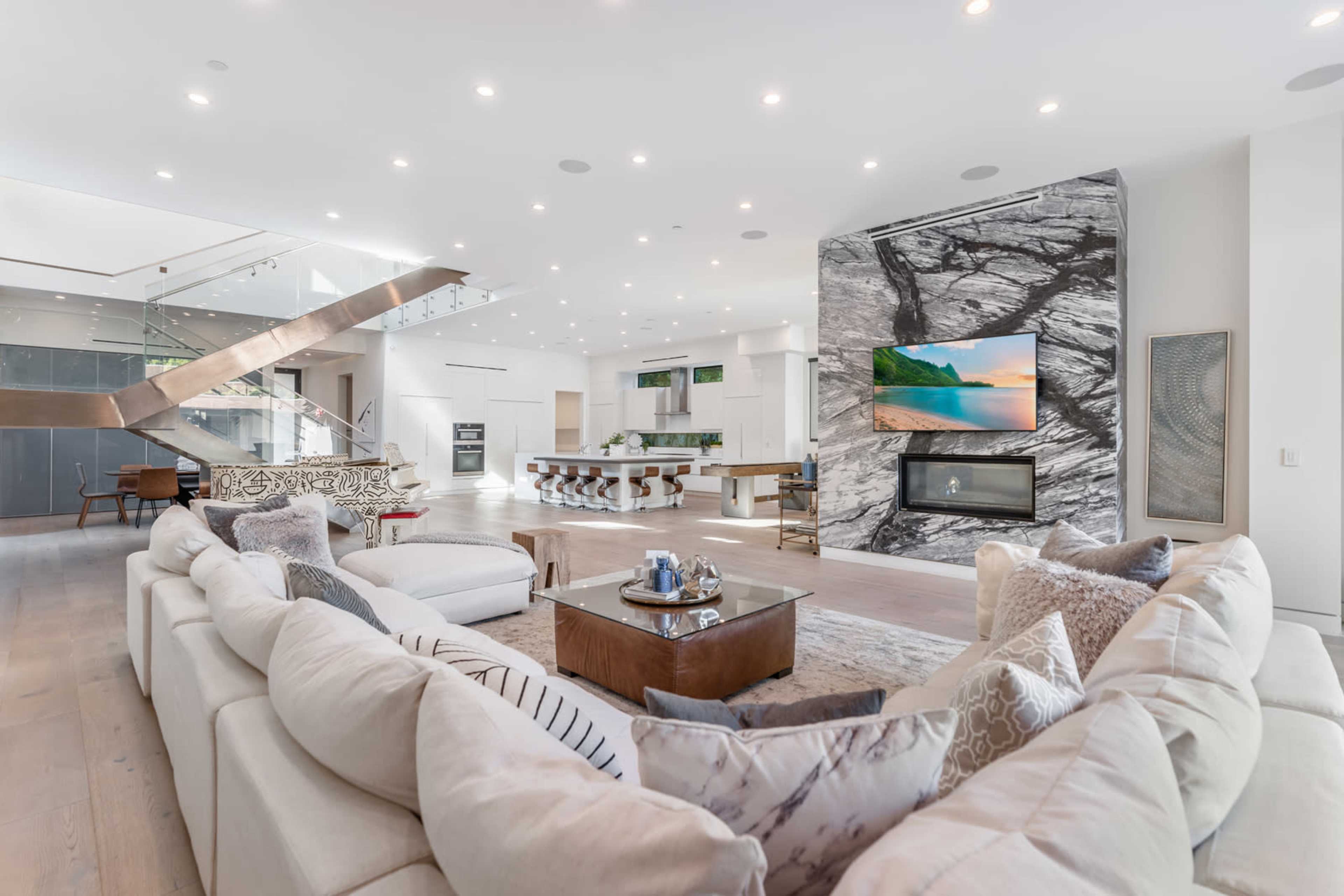 A modern living room featuring a large sectional couch, a coffee table, a sleek stone fireplace, and an open layout with high ceilings and large windows.