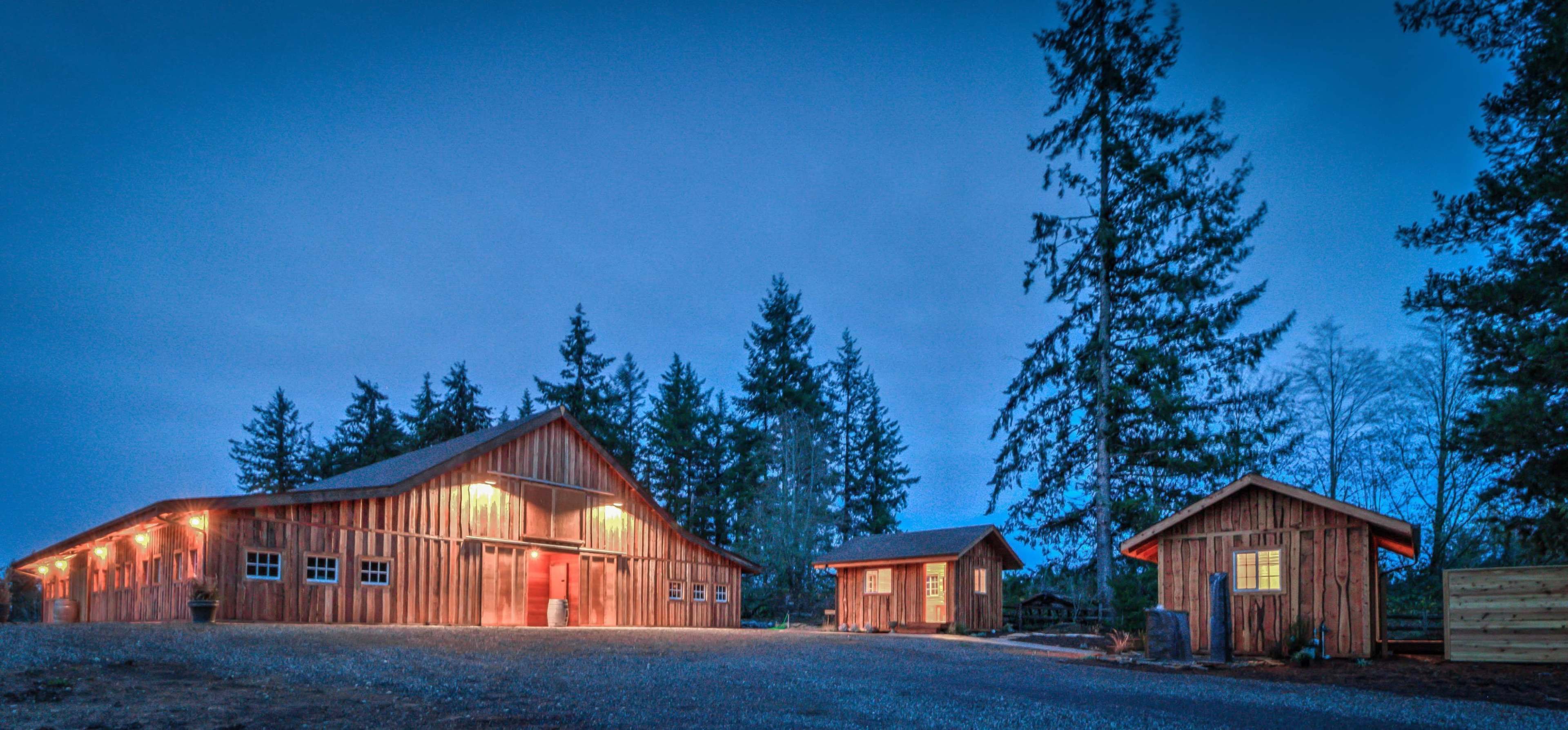 A rustic wooden barn and a smaller outbuilding surrounded by tall trees during twilight.