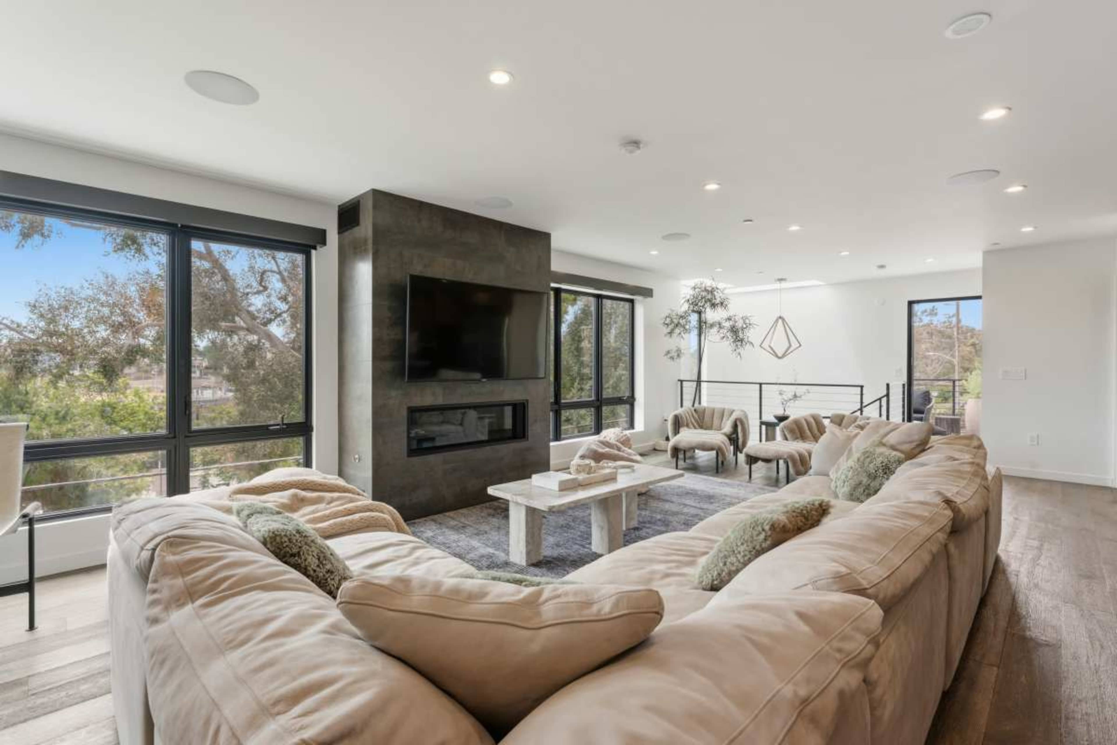 The image shows a modern living room featuring a large sectional sofa, a minimalist coffee table, and large windows that provide natural light and a view of trees outside.