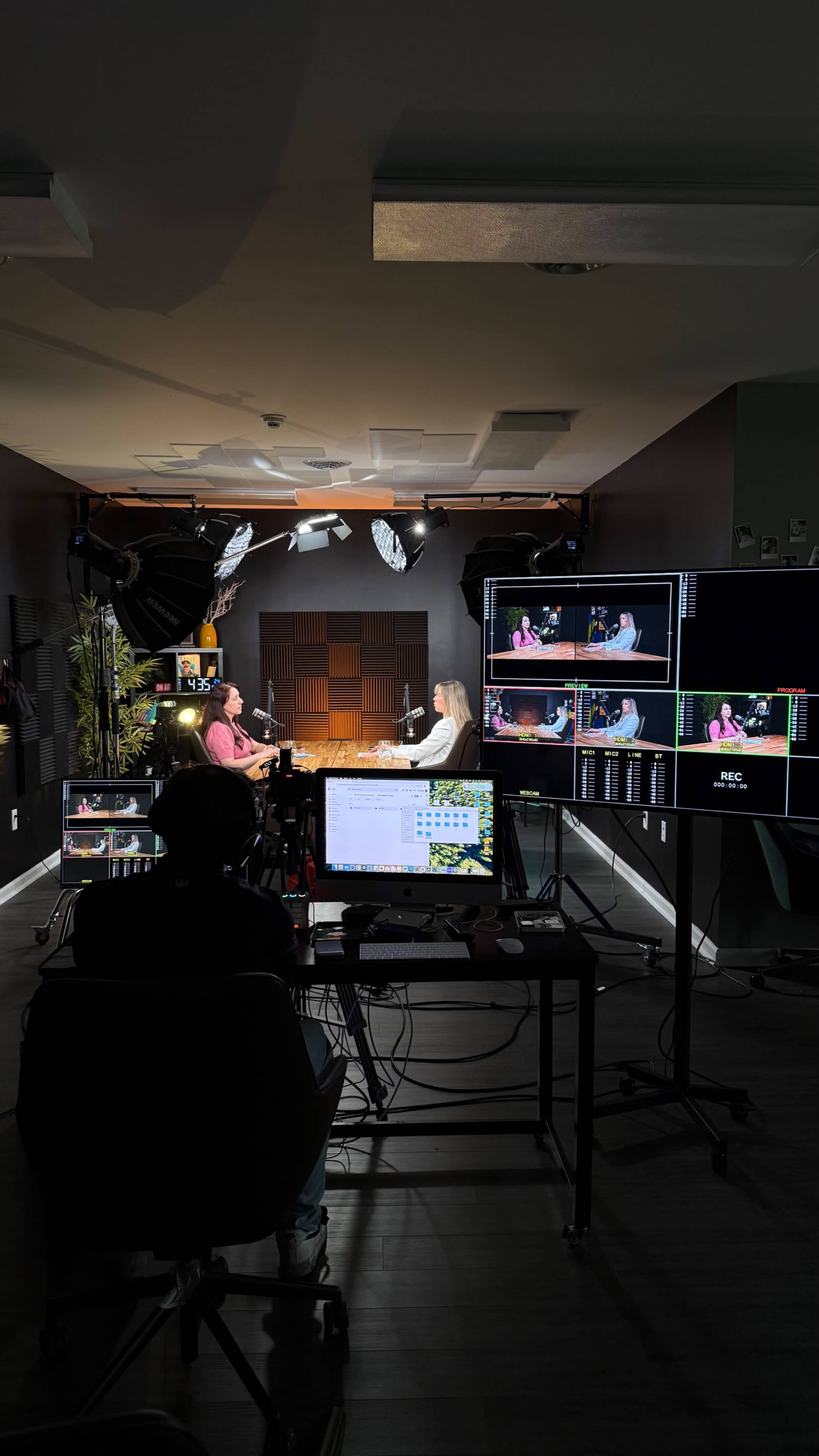 A recording studio setup features two individuals seated at a table, with multiple screens displaying their conversation and various lighting equipment in the background.