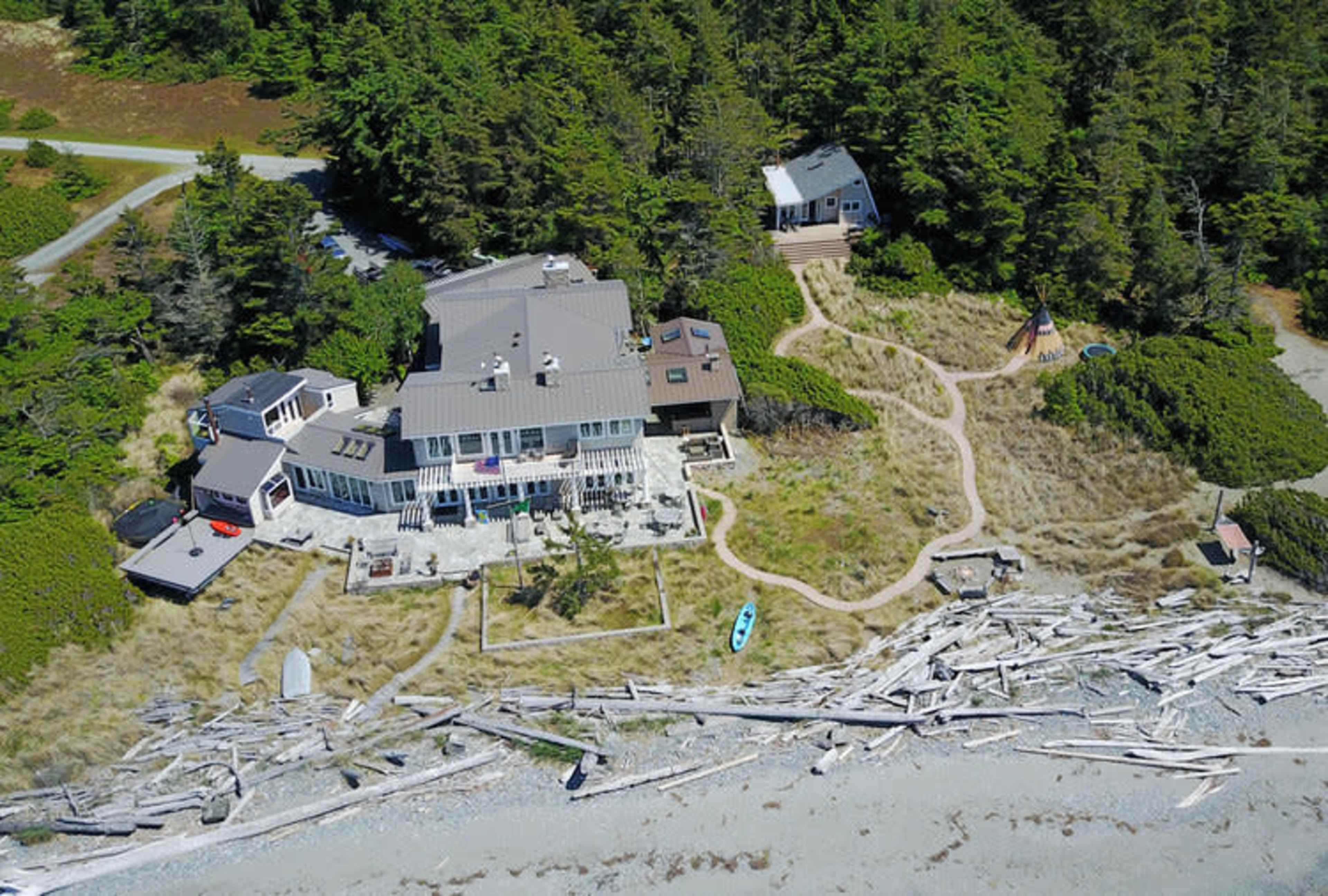 An aerial view of a coastal property featuring a large house, surrounded by trees and a beach with driftwood, along with a path leading to a smaller building.