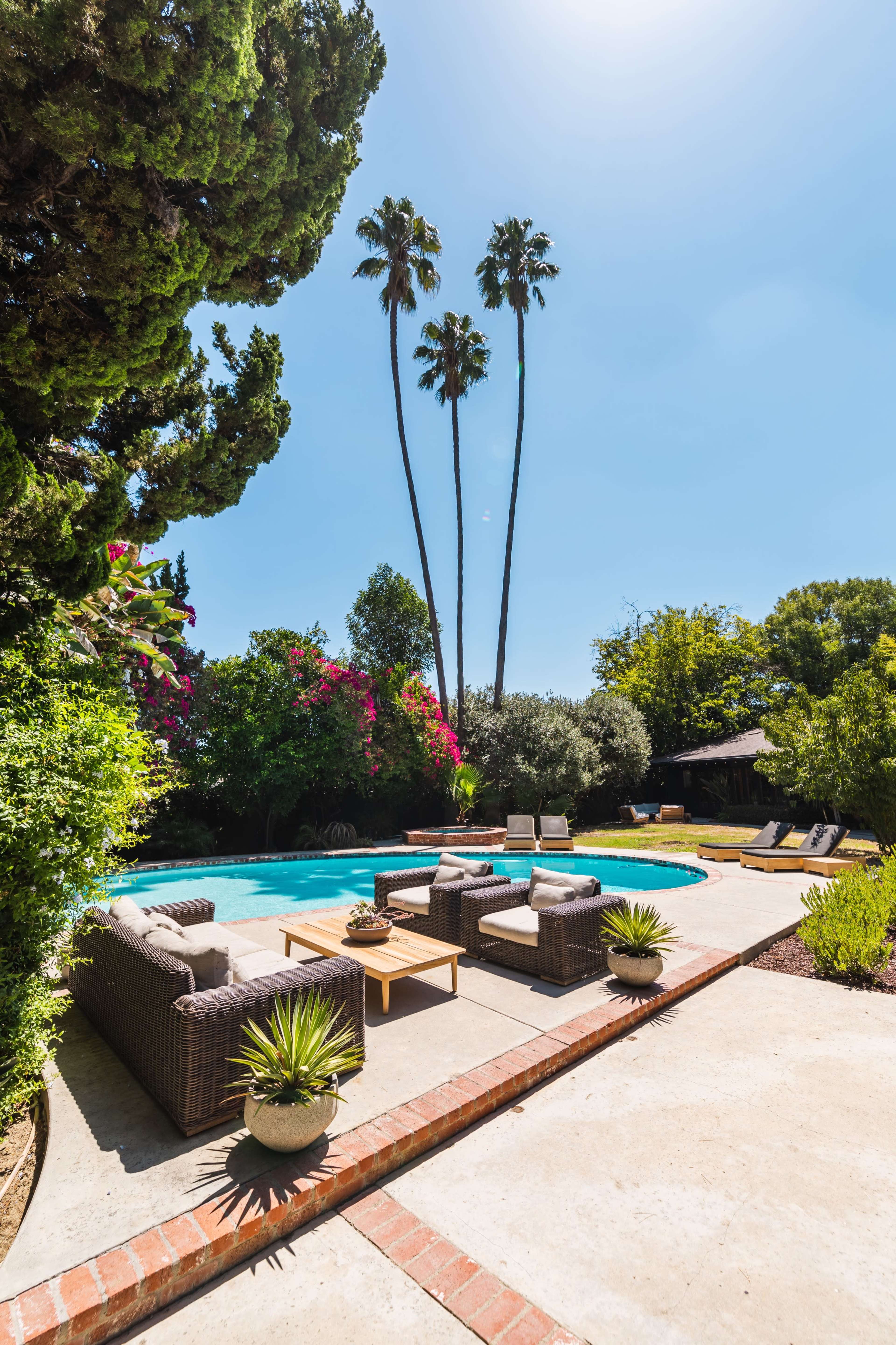 The image shows a backyard featuring a swimming pool surrounded by patio furniture, palm trees, and colorful vegetation.