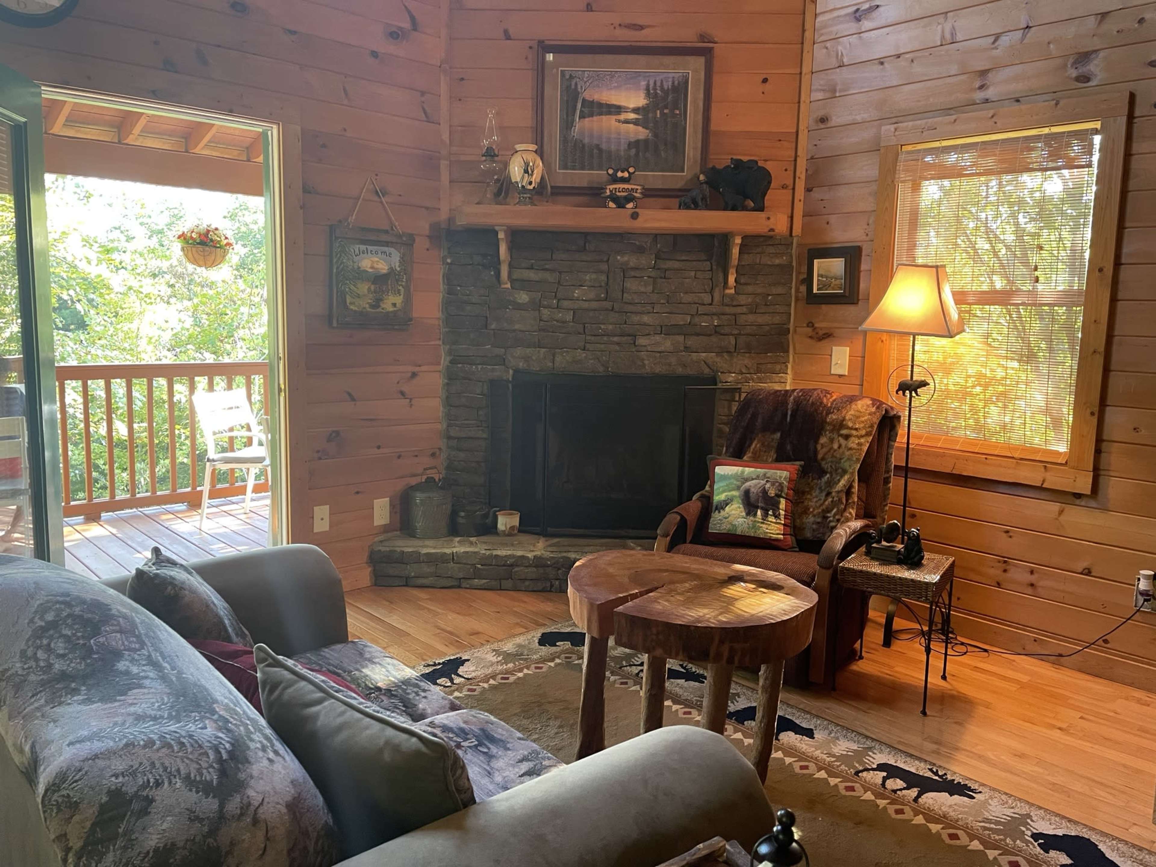 The image shows a cozy wooden cabin interior with a stone fireplace, a comfortable chair, and a small round table beside it, illuminated by natural light from a window.