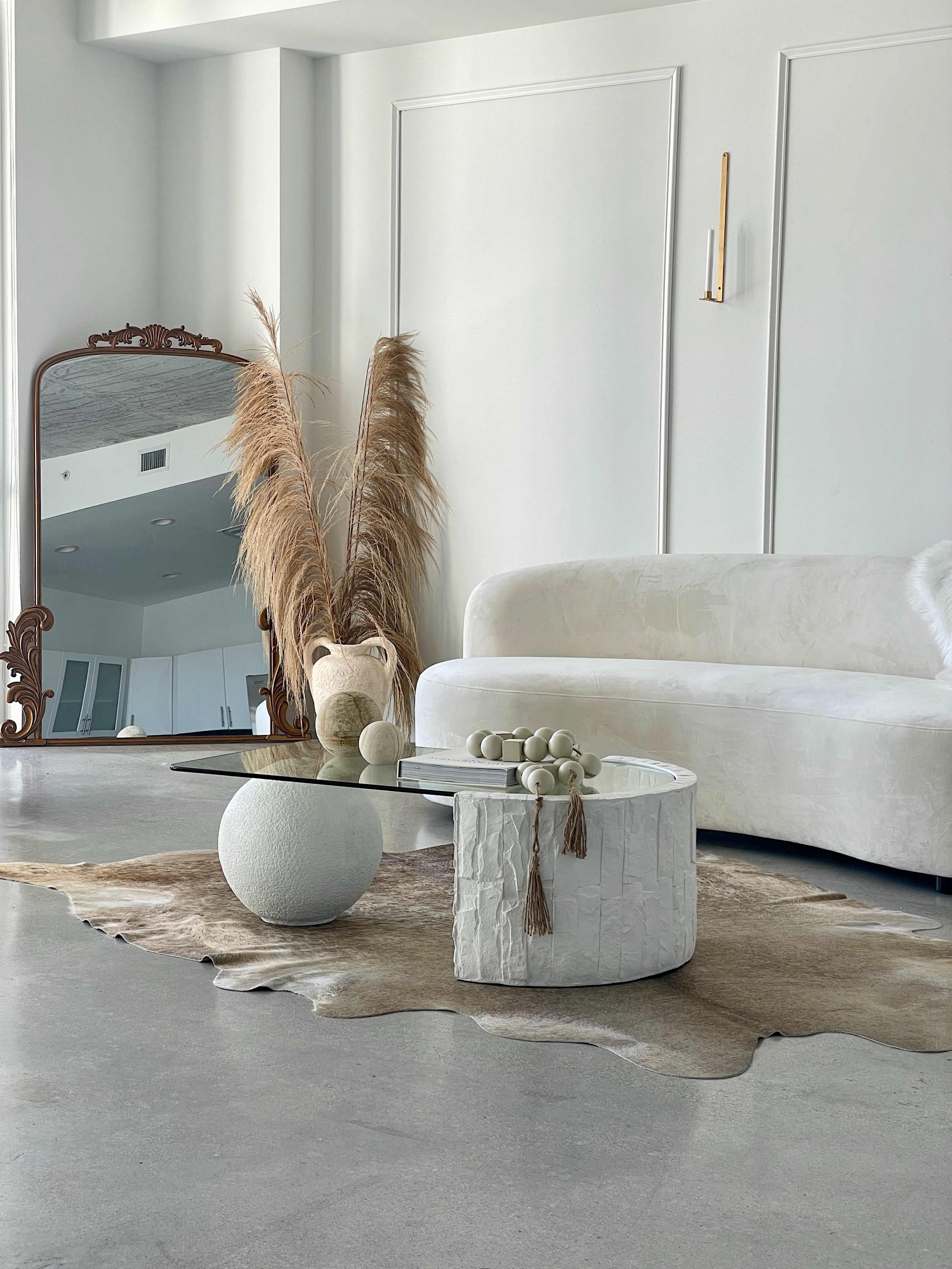 The scene features a minimalist living room with a white curved sofa, a round coffee table, a large mirror, decorative vases, and dried plants on a textured rug.