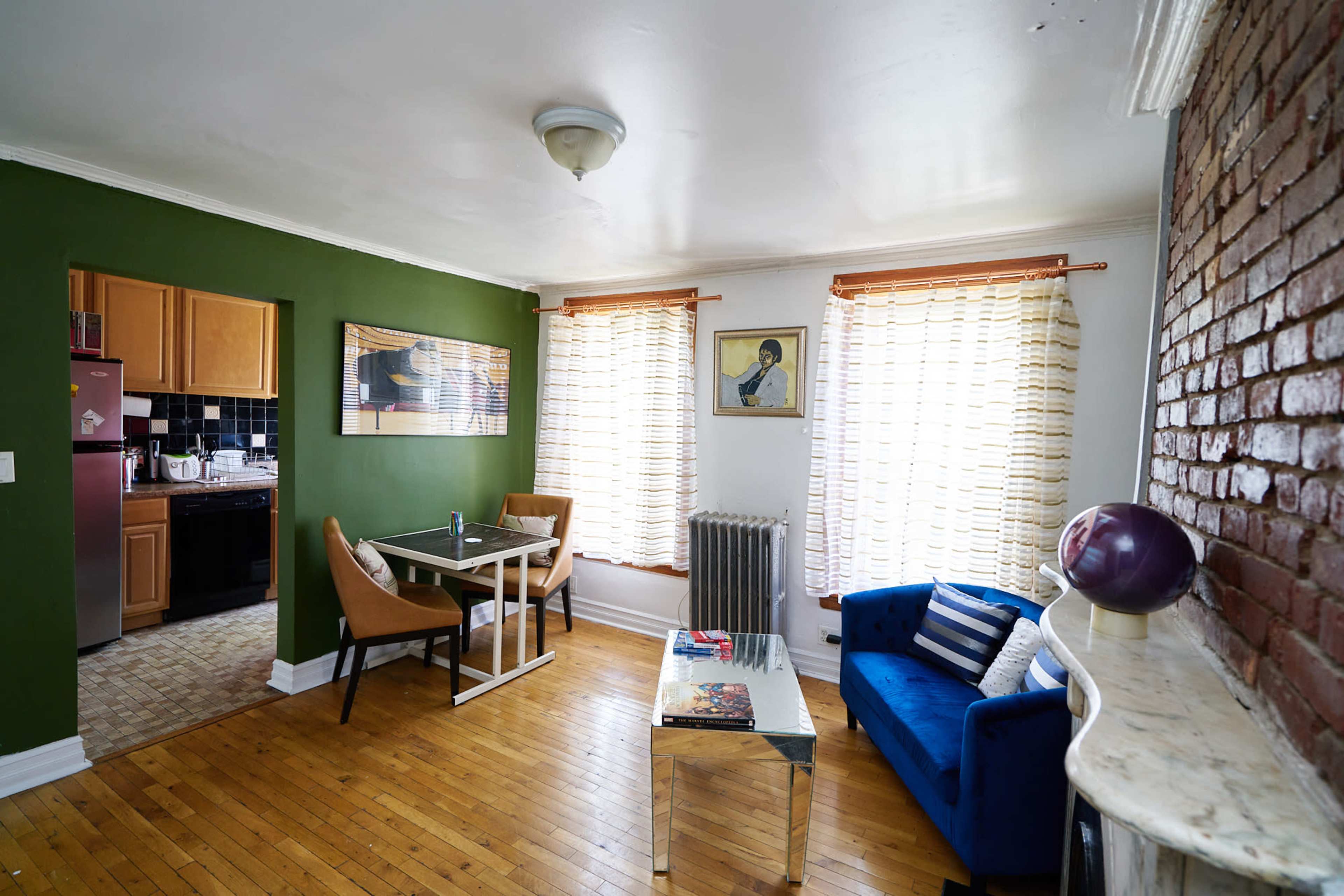 The image shows a living space with a blue sofa, a glass coffee table, a dining area, and windows dressed in light curtains, adjacent to a kitchen with dark cabinets.