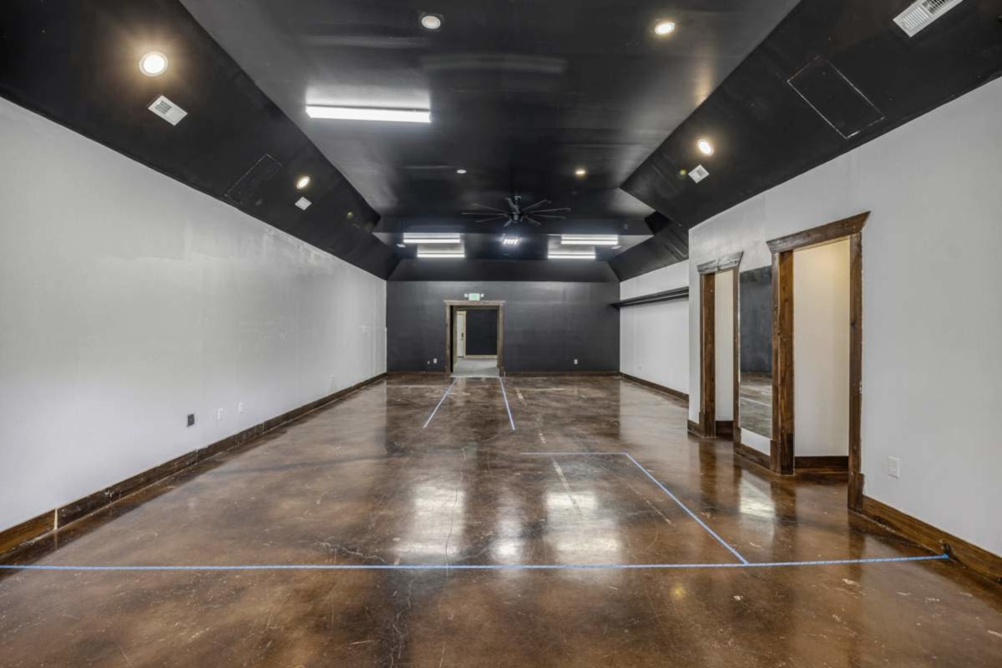 The image shows an empty, spacious room with a polished concrete floor, dark ceiling, and white walls, featuring recessed lighting and a central ceiling fan.