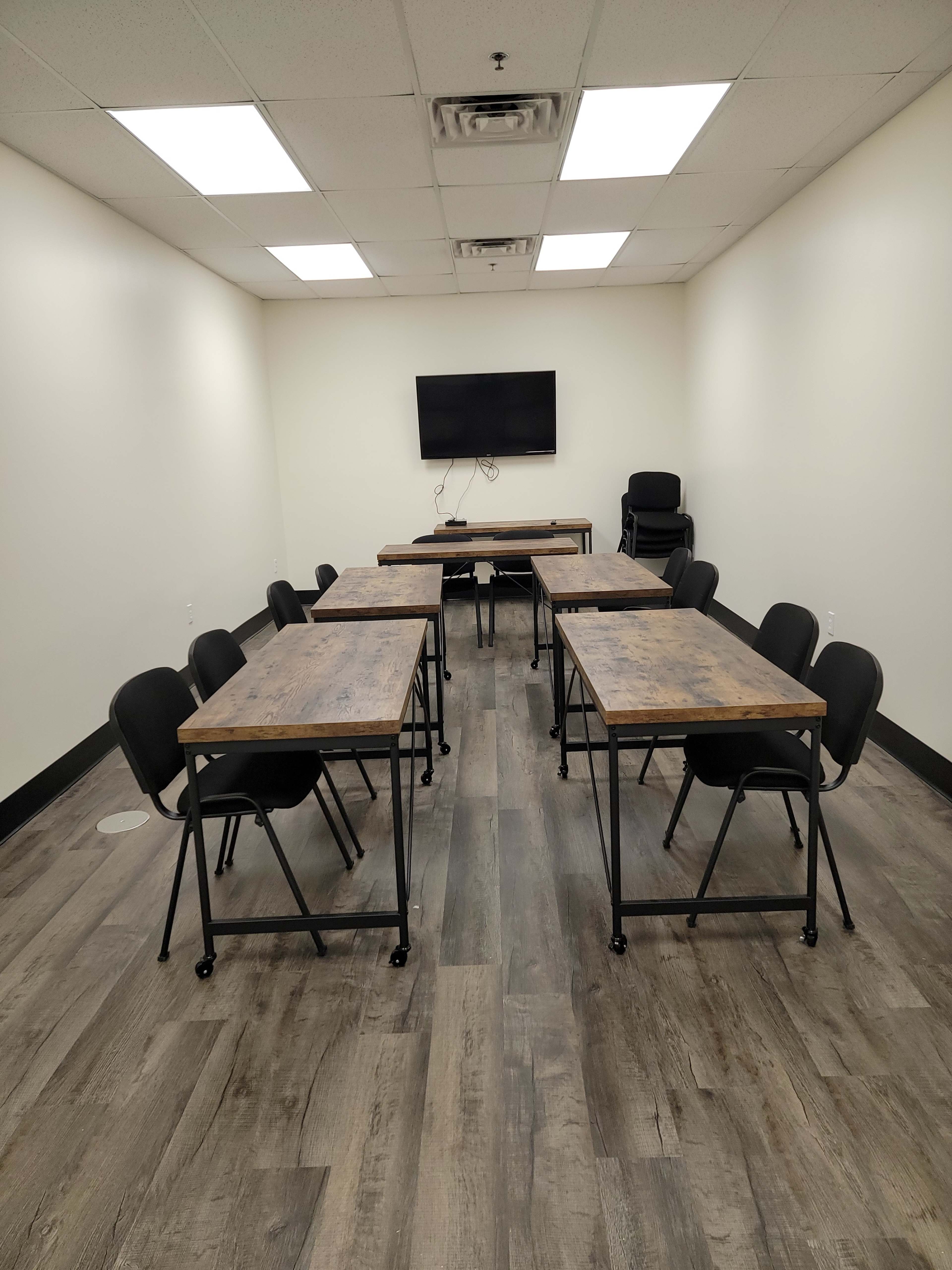 A meeting room is arranged with several wooden tables and black chairs, featuring a wall-mounted television.