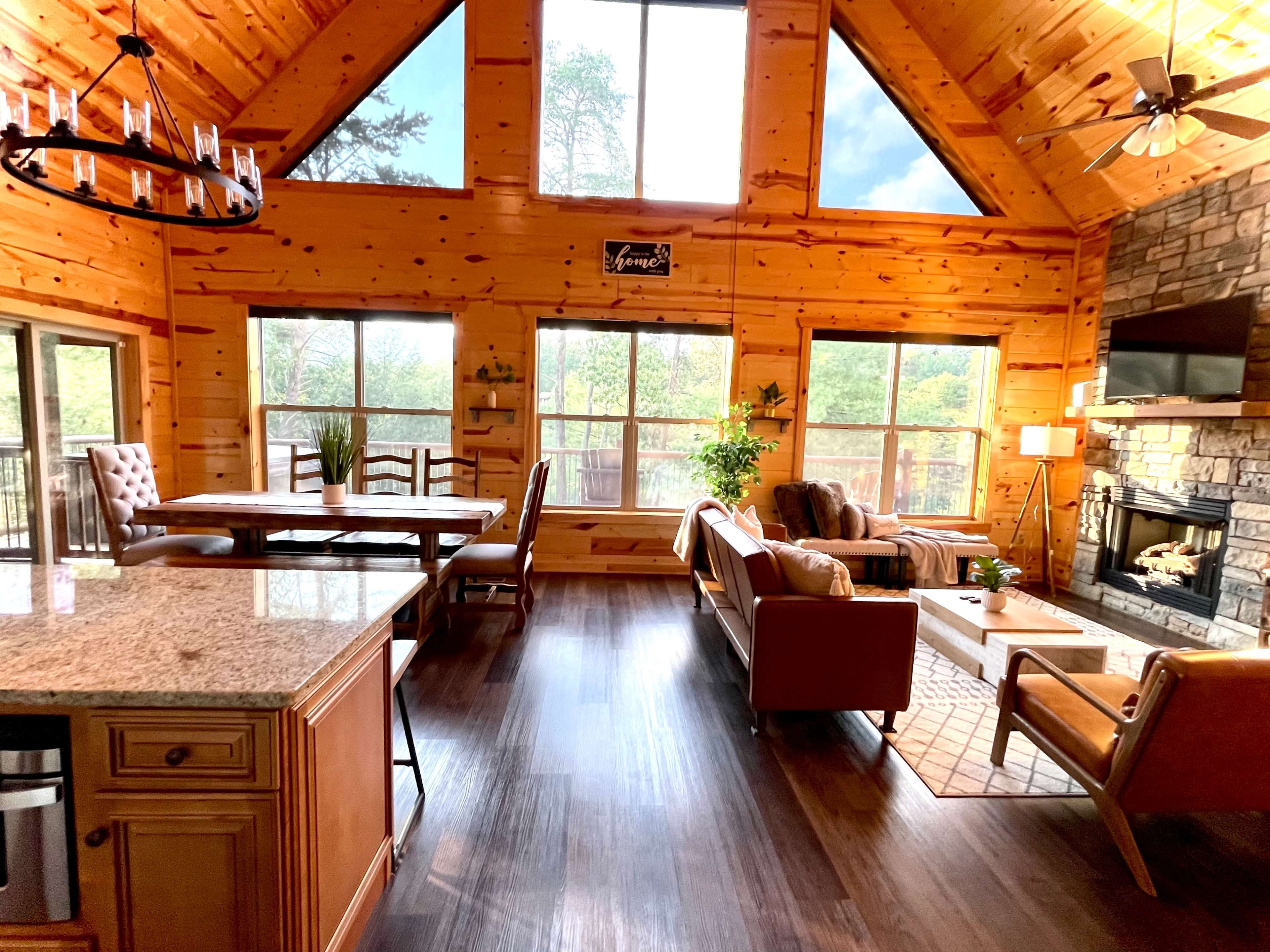 The interior of a wooden cabin features a living room with large windows, a stone fireplace, and a dining area with a wooden table.