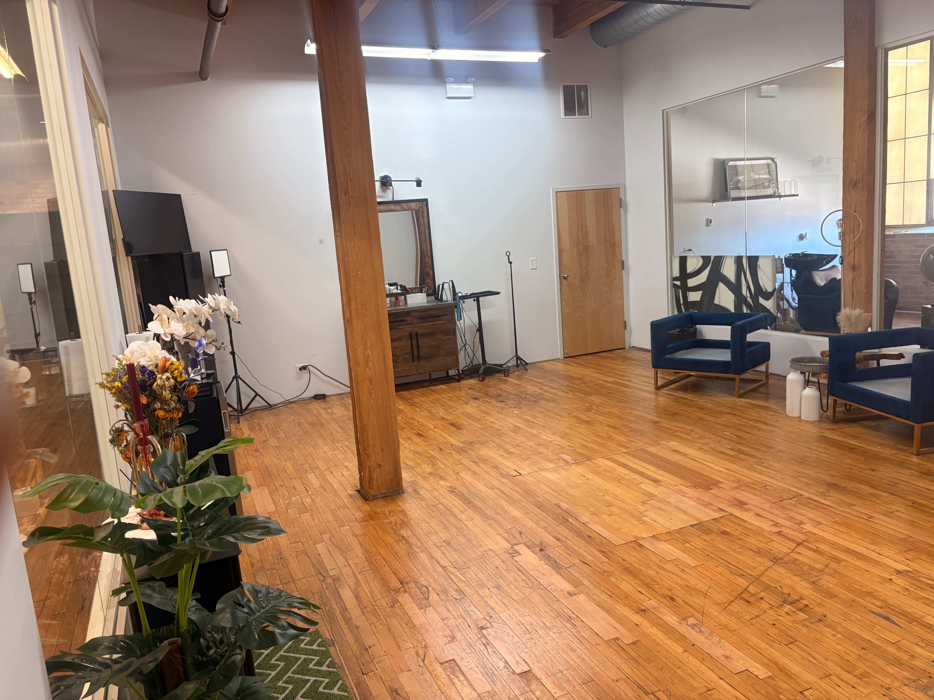 The image shows a spacious, well-lit room with wooden flooring, featuring a few potted plants, two blue armchairs, and wooden beams supporting the ceiling.