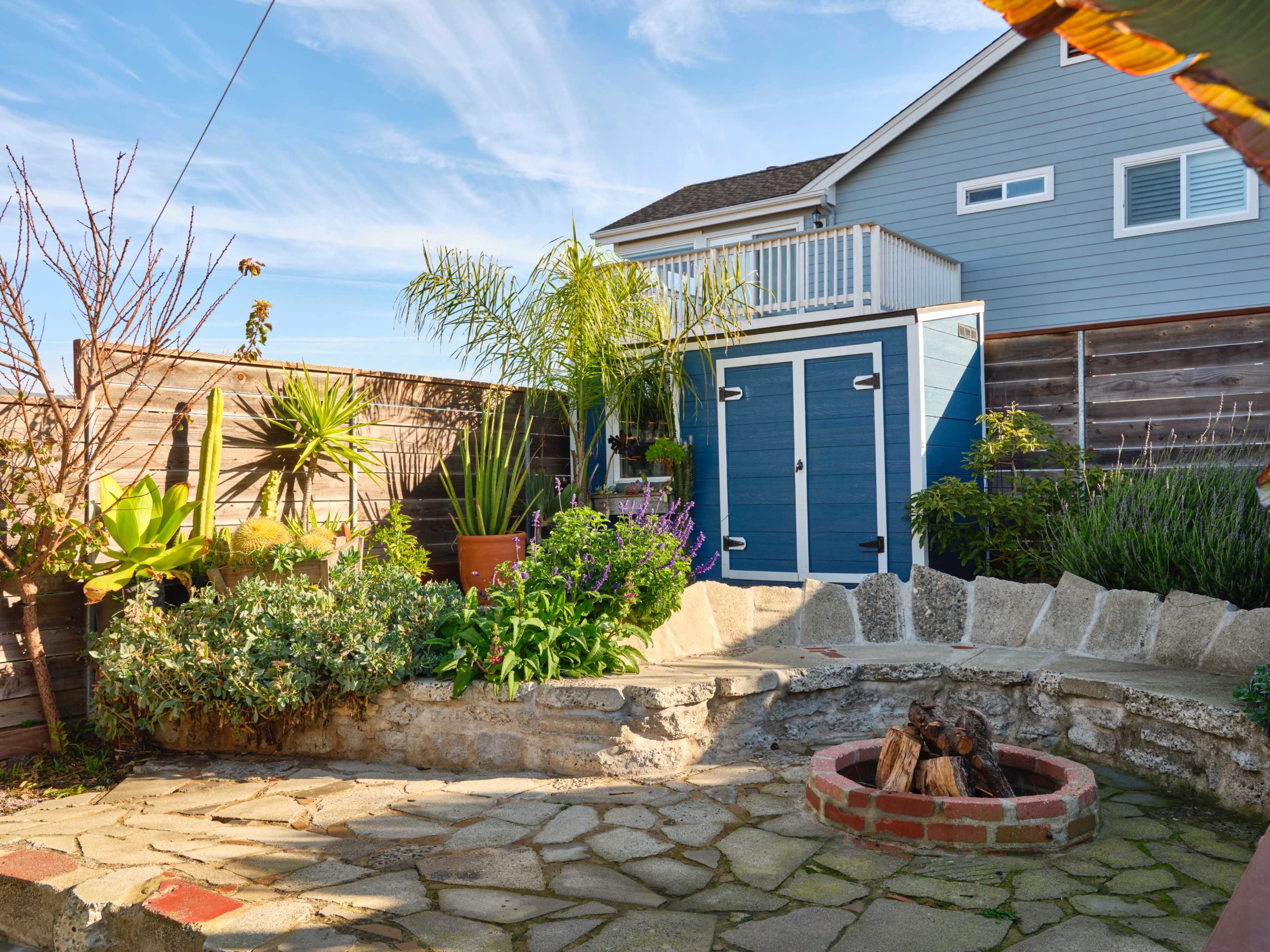 A landscaped backyard featuring a stone patio, a fire pit, various plants, and a blue shed beside a house with a balcony.