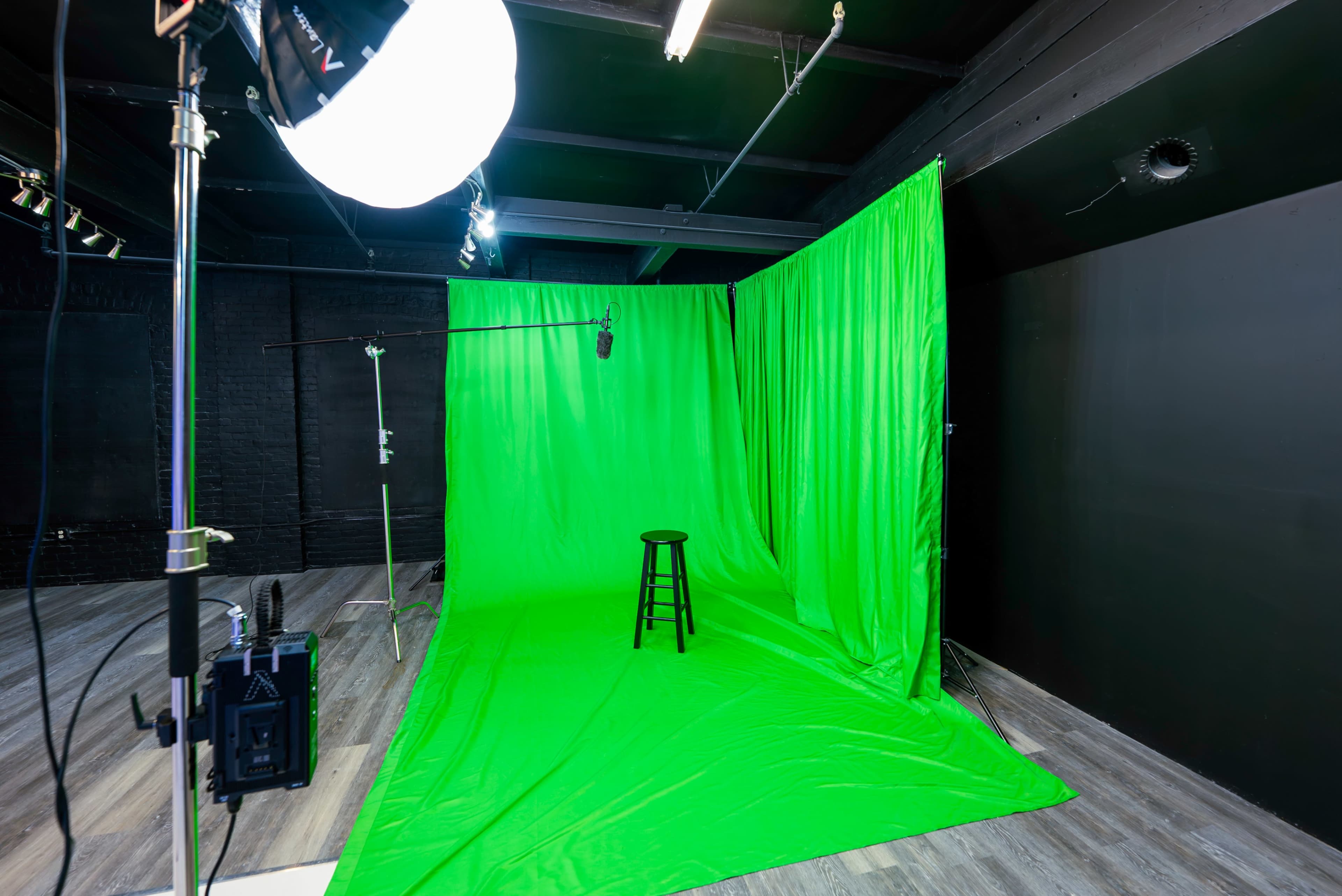 The image shows a studio setup with a green screen backdrop, a black stool in front of it, and lighting equipment positioned off to the side.