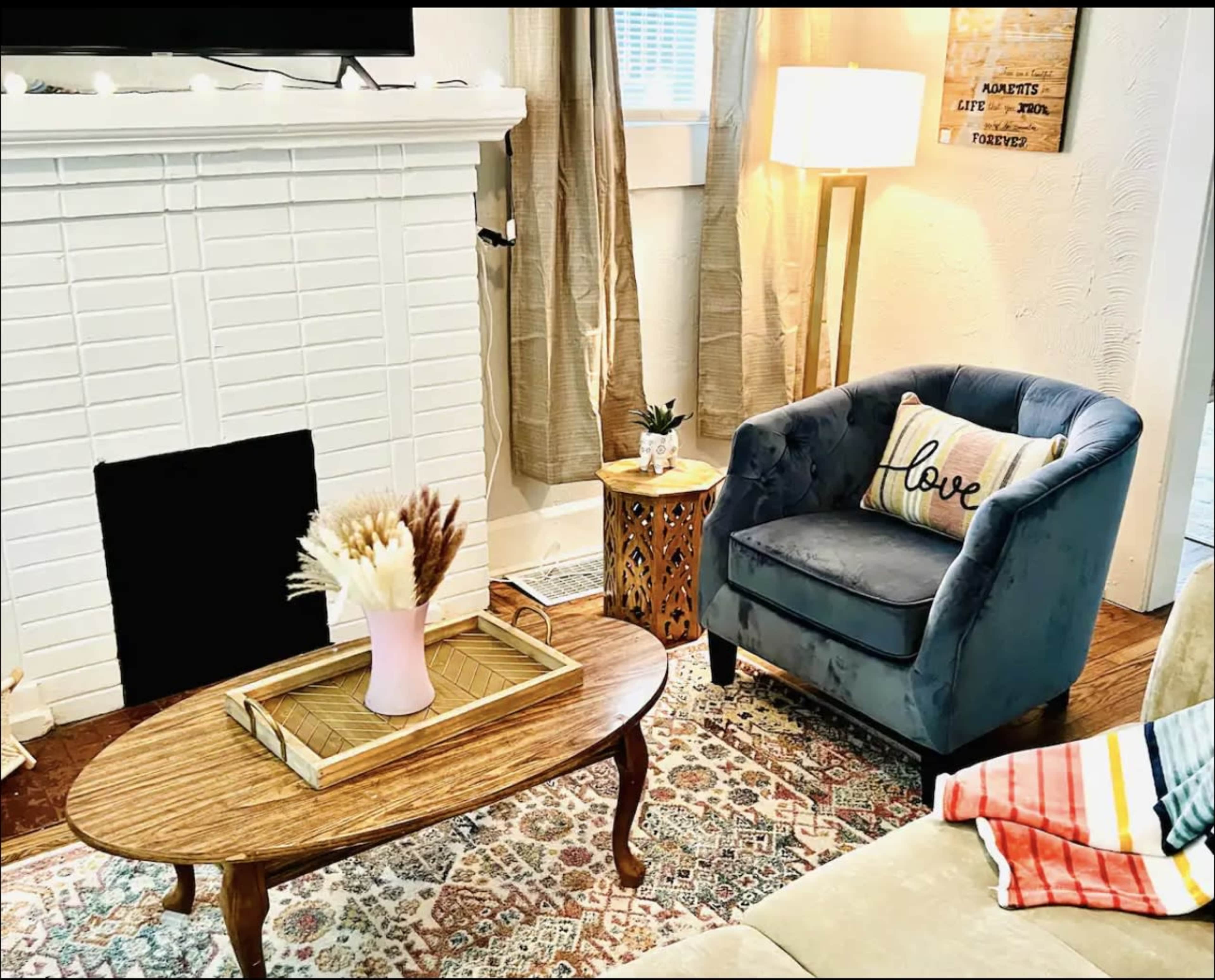 The image shows a cozy living room featuring a blue armchair with a "love" pillow, a wooden coffee table with a tray and a vase of dried flowers, and a patterned area rug.