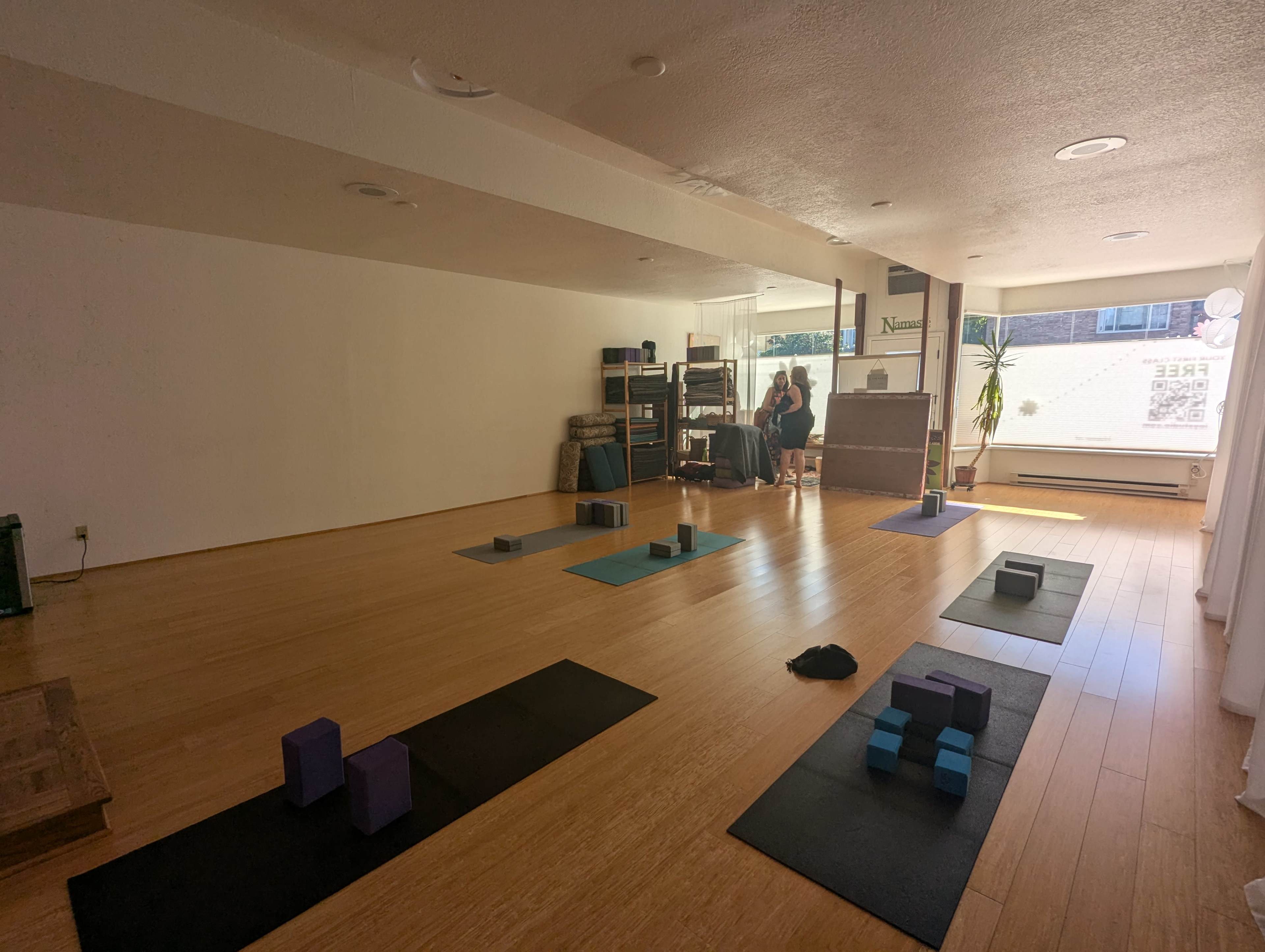 A yoga studio features wooden flooring with exercise mats, blocks, and props arranged in a spacious layout.