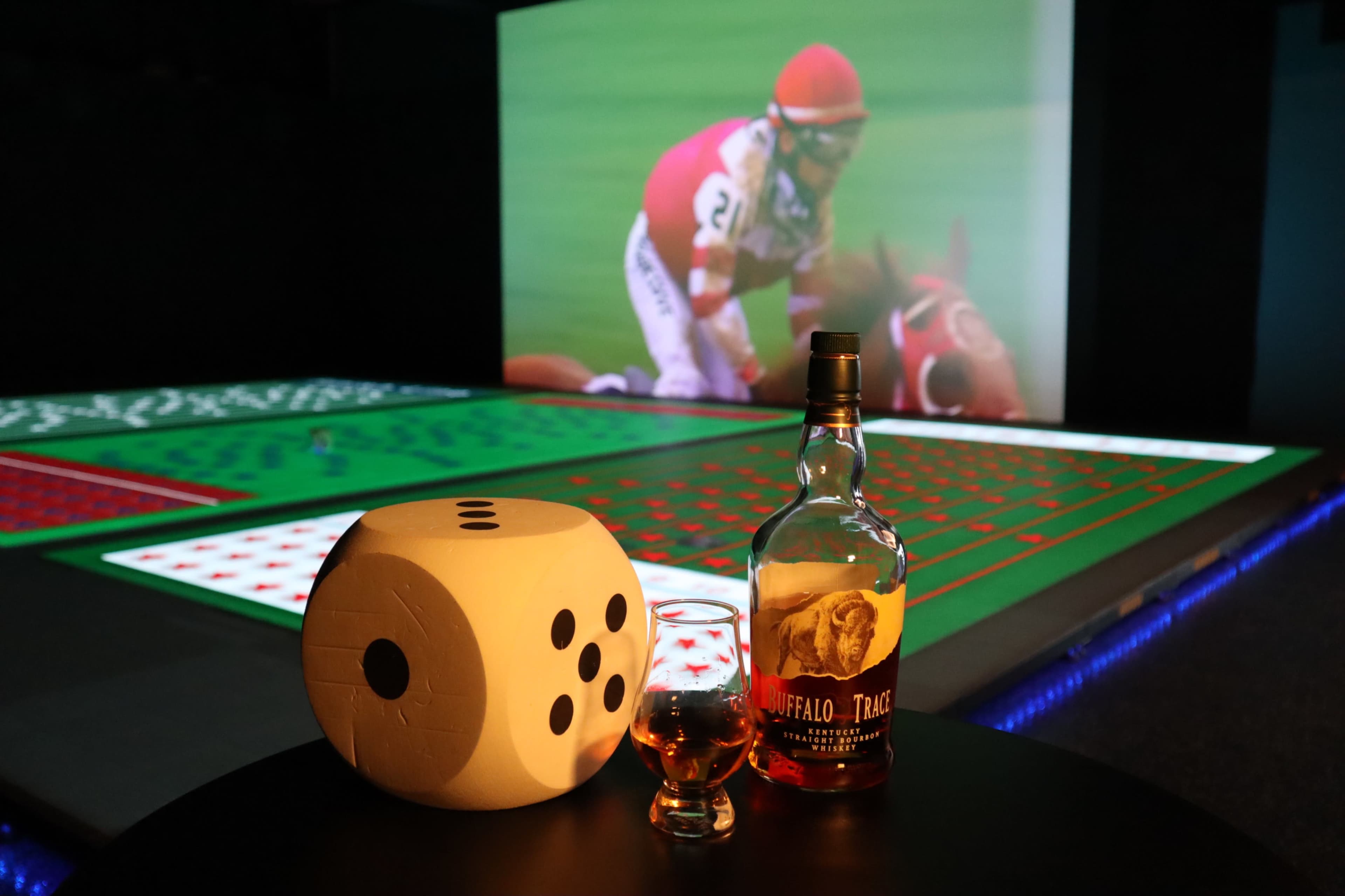 A large die, a glass of whiskey, and a bottle of bourbon are positioned in front of a digital screen displaying a sports event.