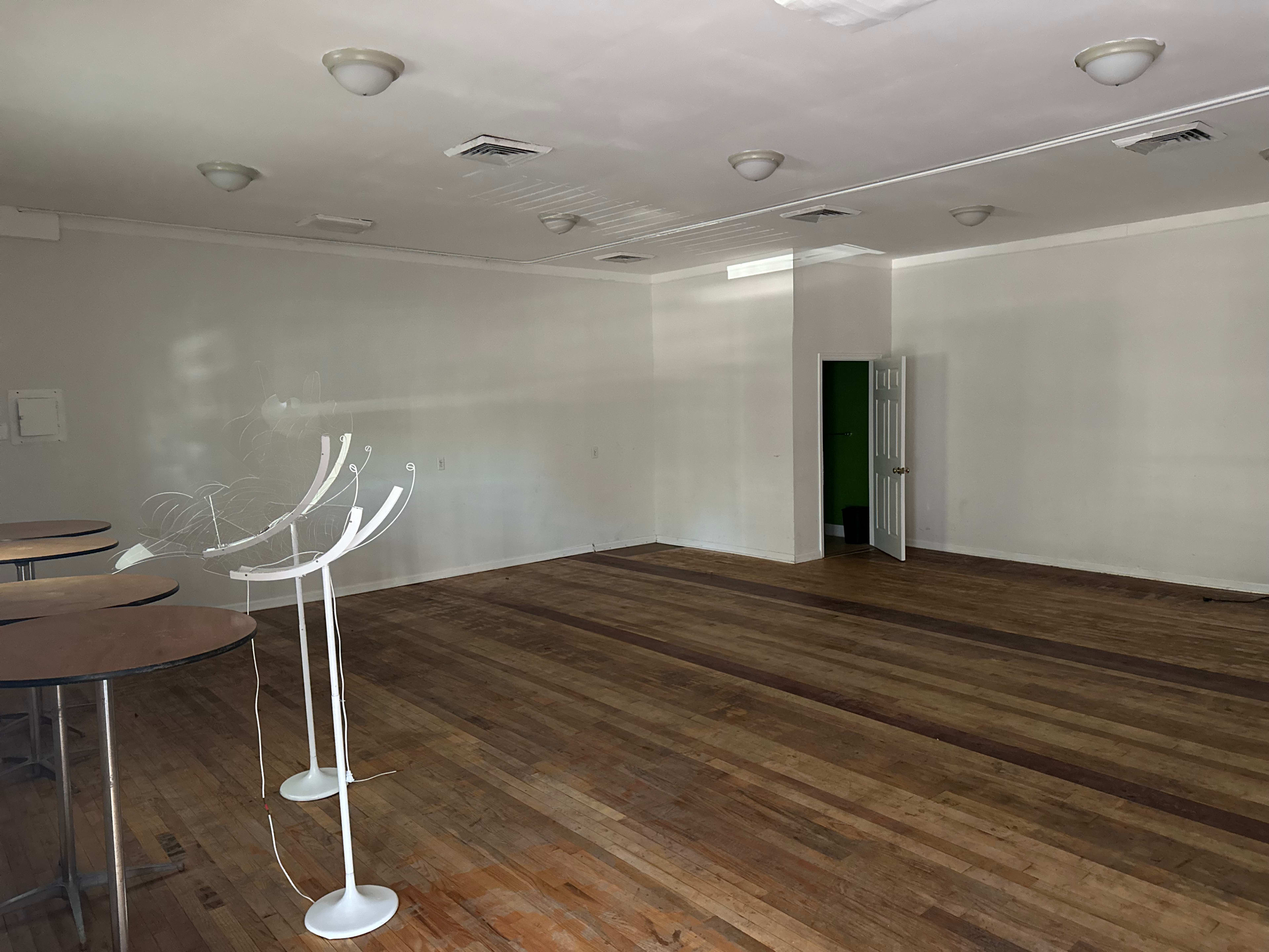 An empty room with wooden flooring, featuring a few tables and a door leading to a green-walled space.