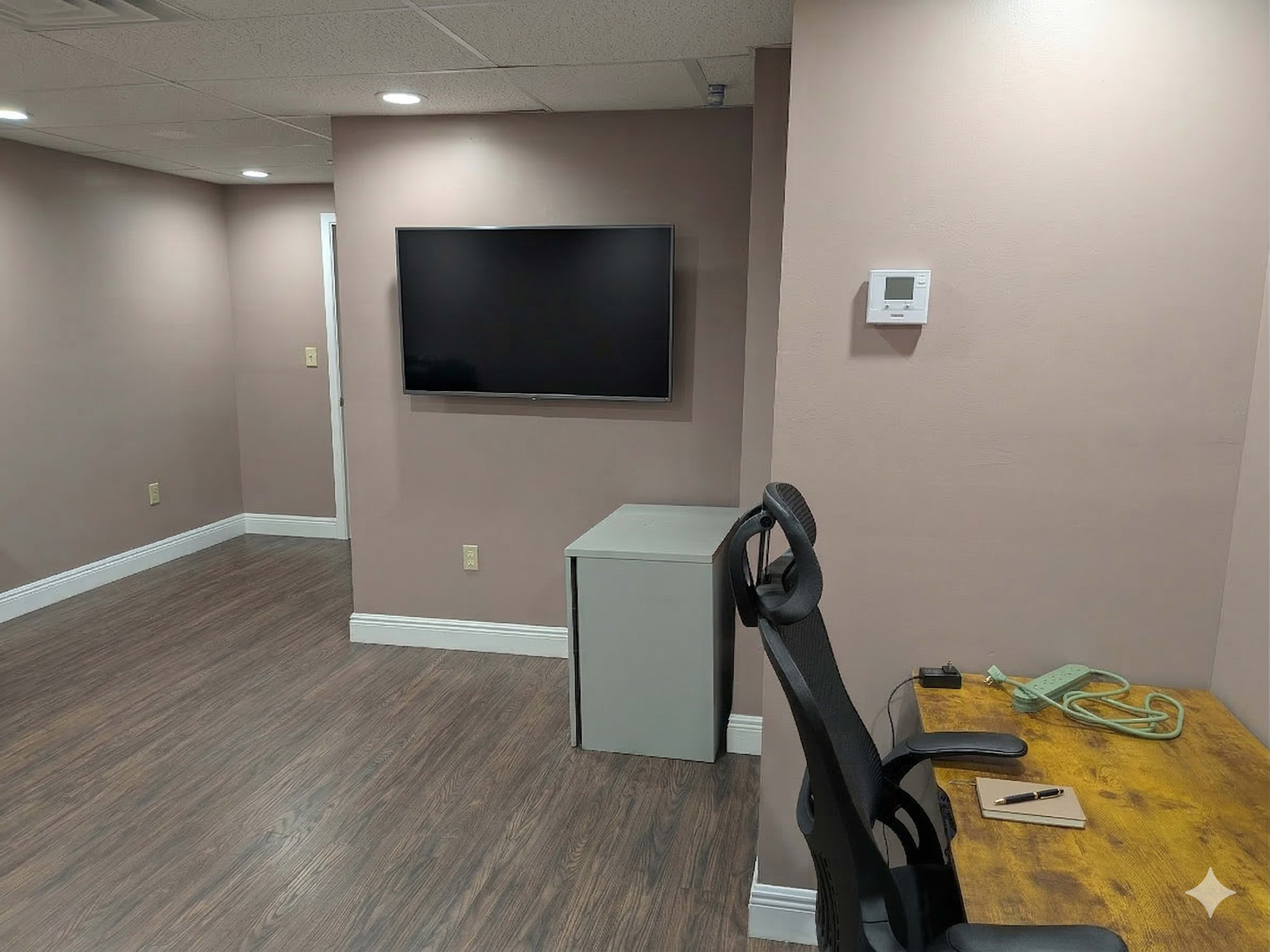 The image shows a small office space with a television mounted on the wall, a gray cabinet, a wooden desk with an office chair, and light-colored walls.