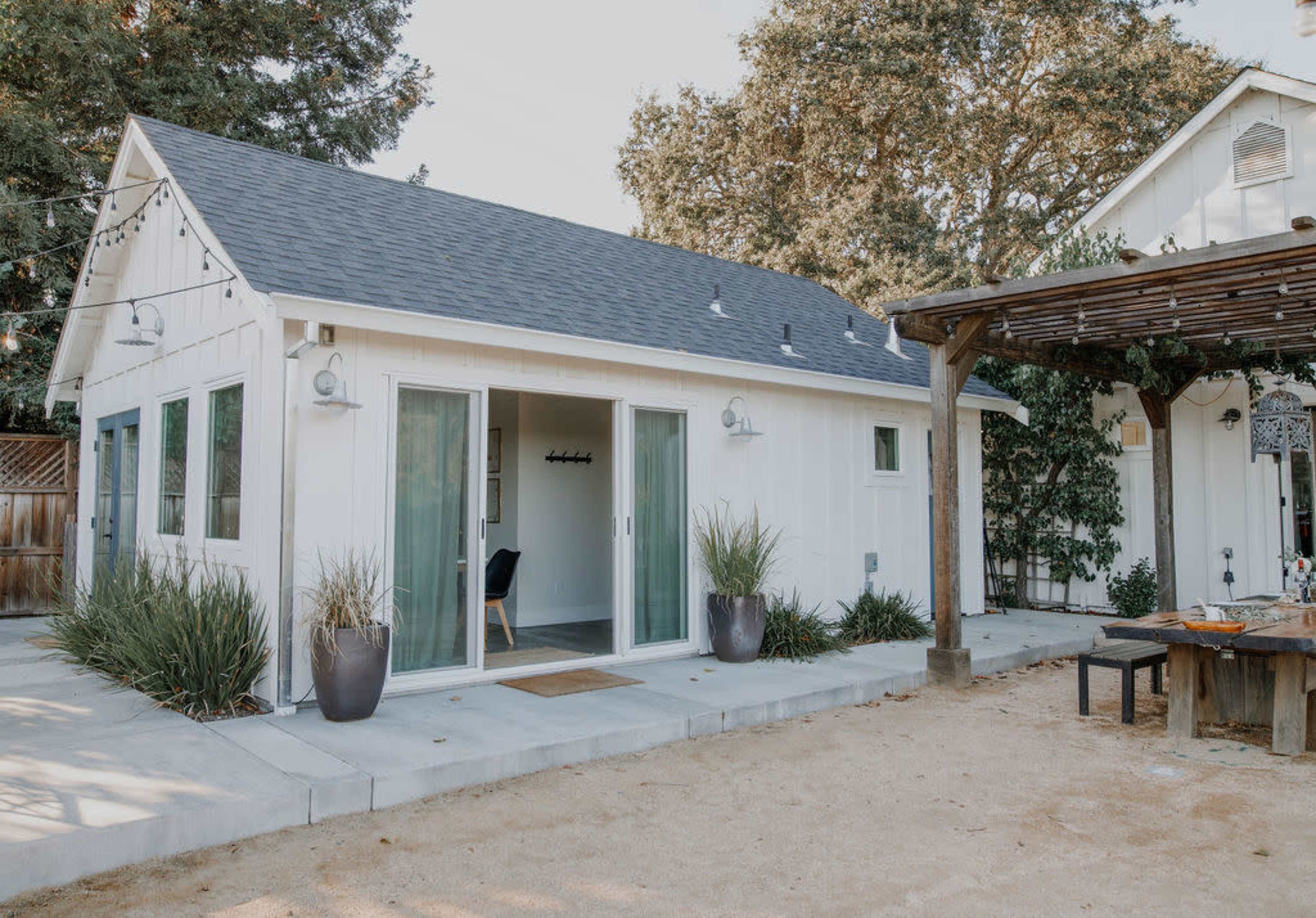 A modern, white cottage with large glass doors opens onto a patio area, surrounded by greenery and a shaded pergola.