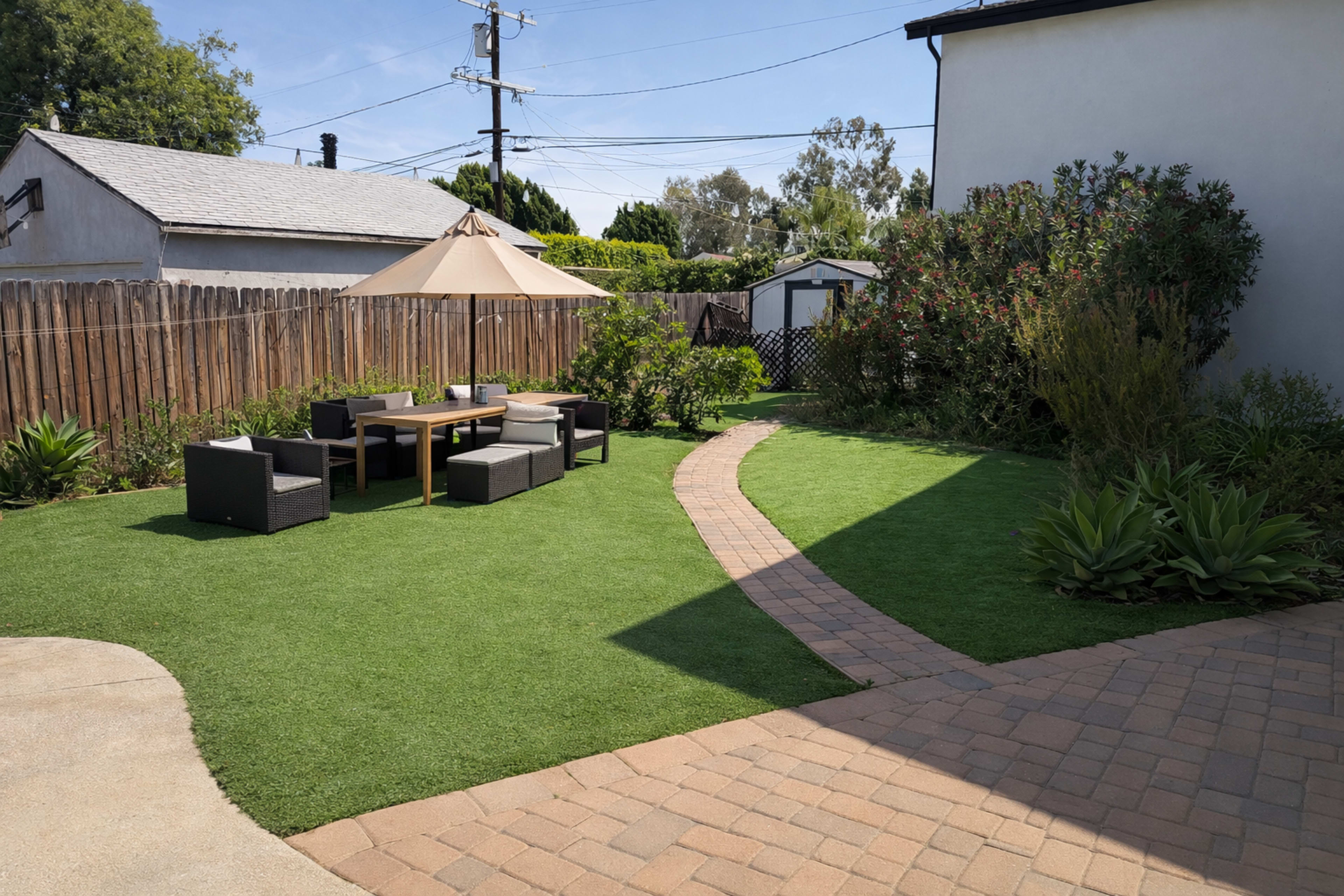 The image shows a landscaped backyard featuring a paved path, a wooden table with chairs under an umbrella, and a lawn area with plants and bushes.