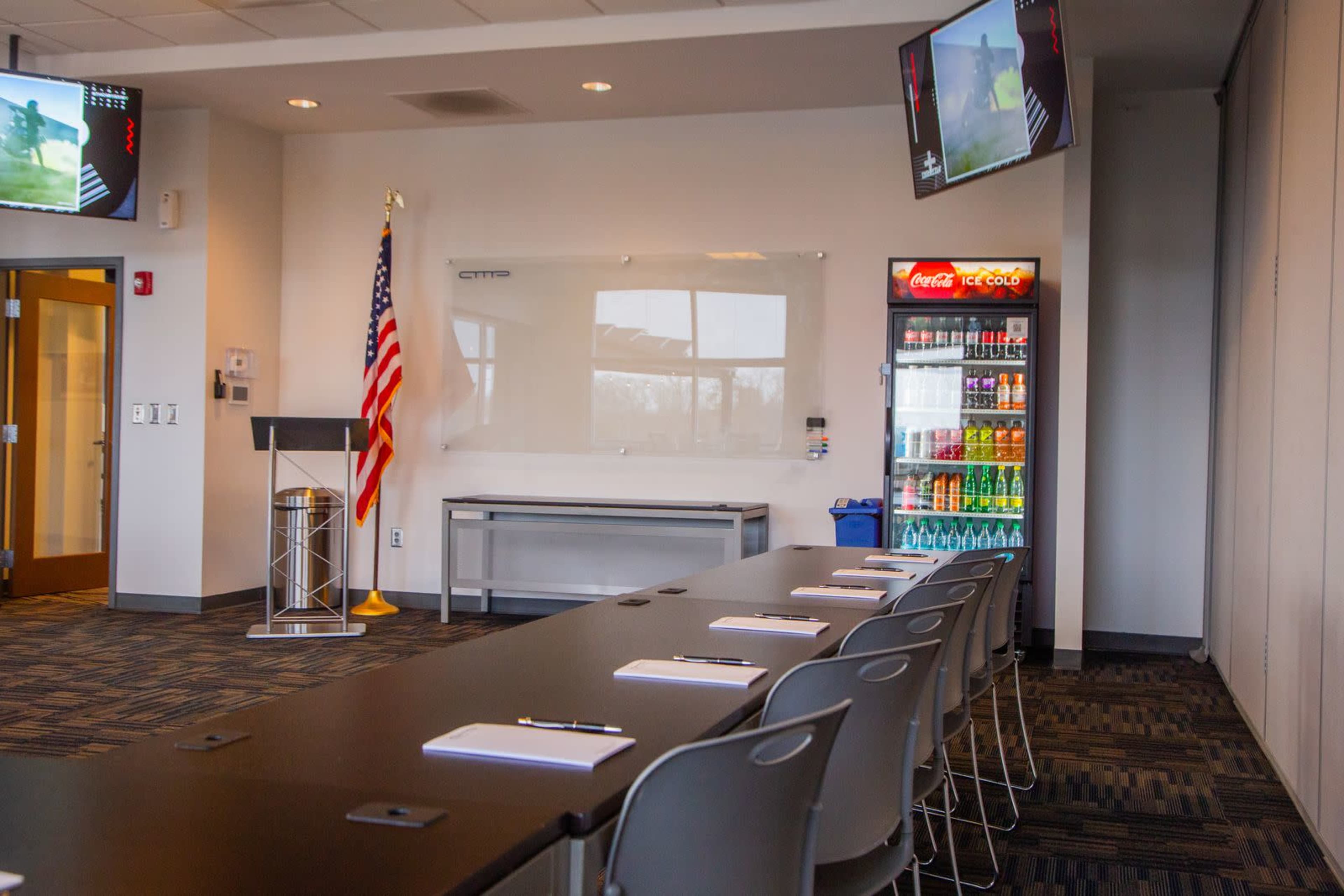 A modern conference room features a long table with seating, a podium, an American flag, two screens displaying images, and a beverage fridge.