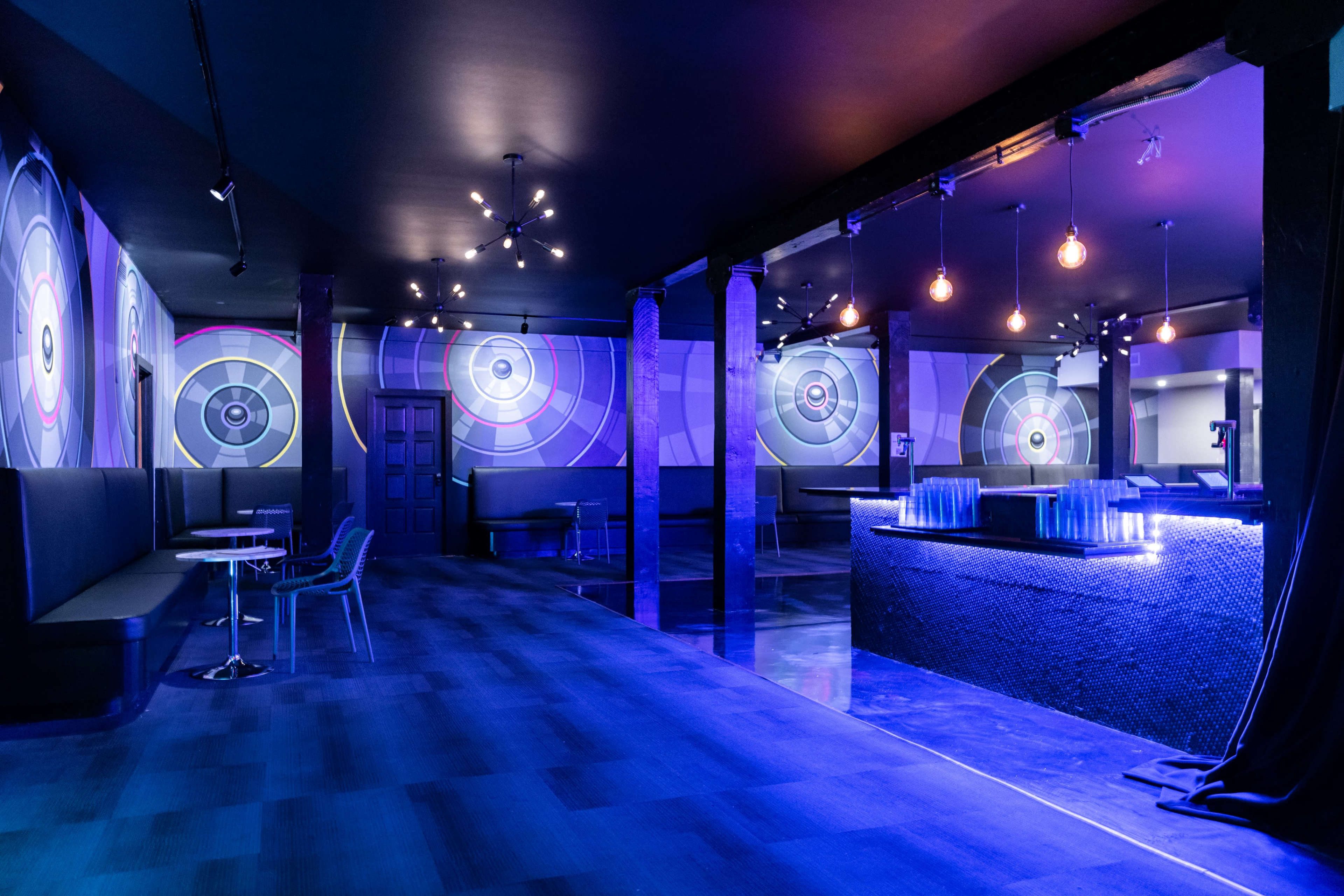 The image shows a modern lounge decorated with large circular wall graphics, dim lighting, and a bar area with illuminated features.