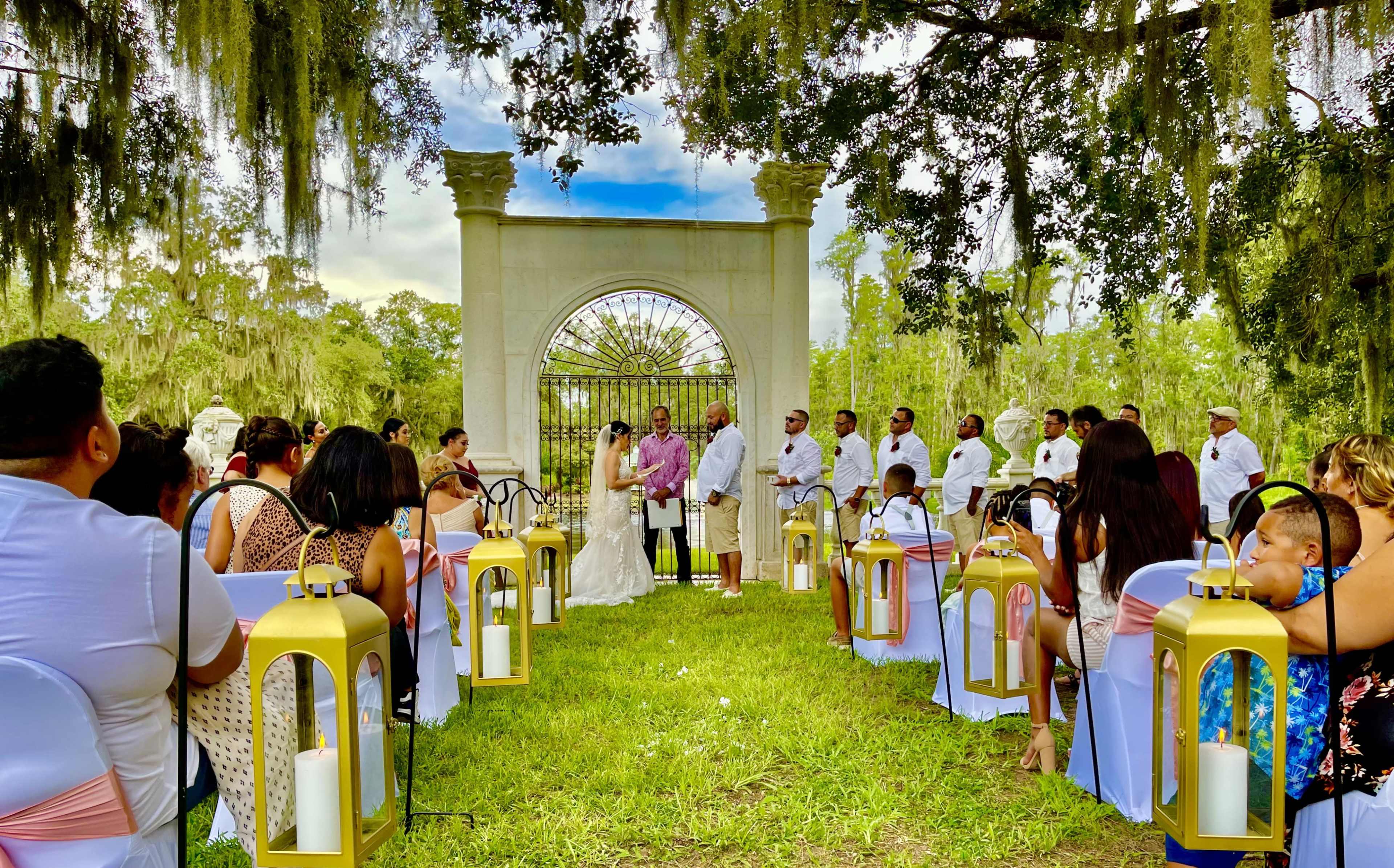 A wedding ceremony takes place outdoors under large trees, with guests seated on chairs aligned in two rows facing the couple and an archway in the background.