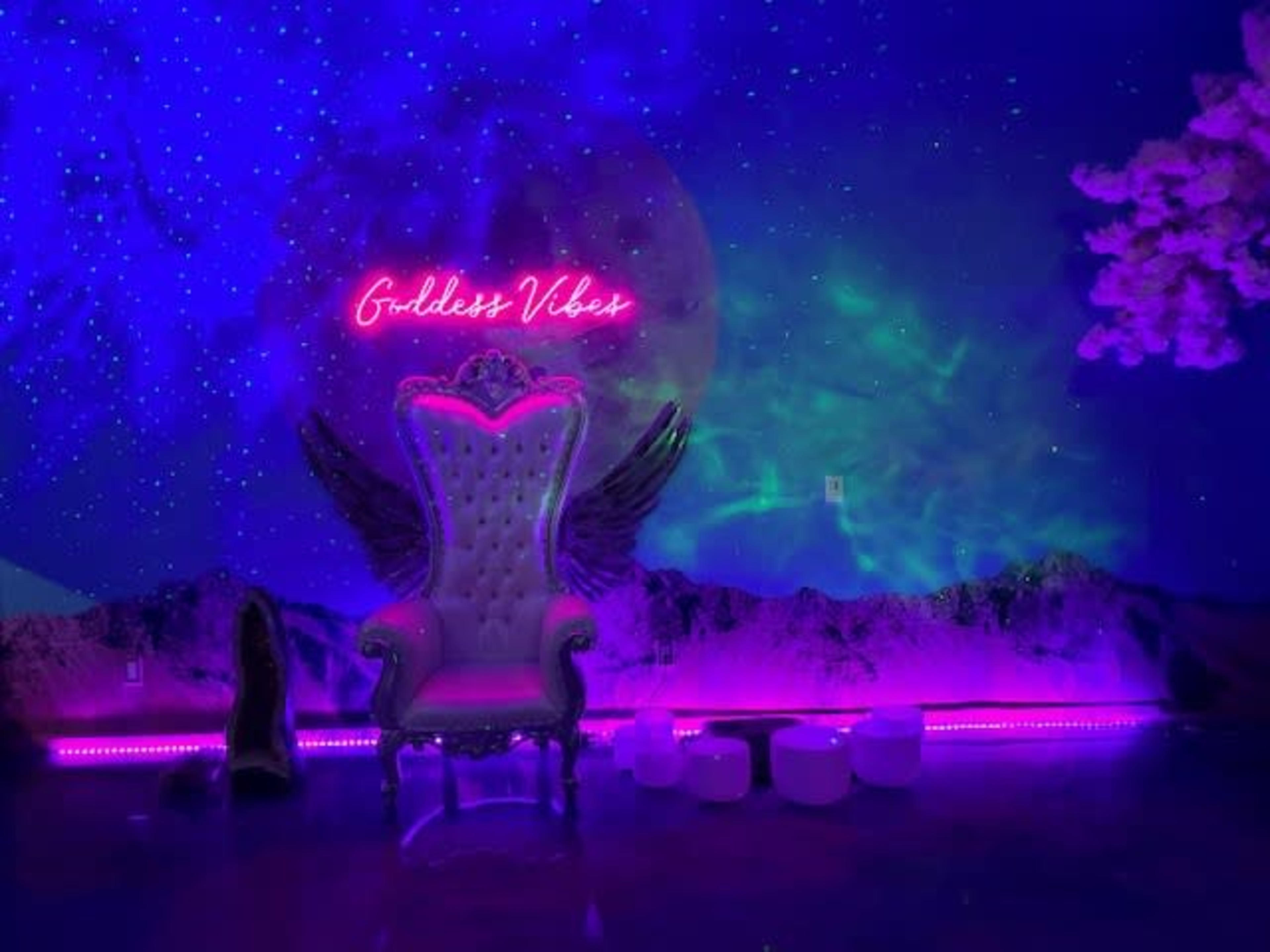 A lavish throne with decorative wings sits in front of a vibrant, colorful mural featuring a cosmic scene and the neon phrase "Goddess Vibes."