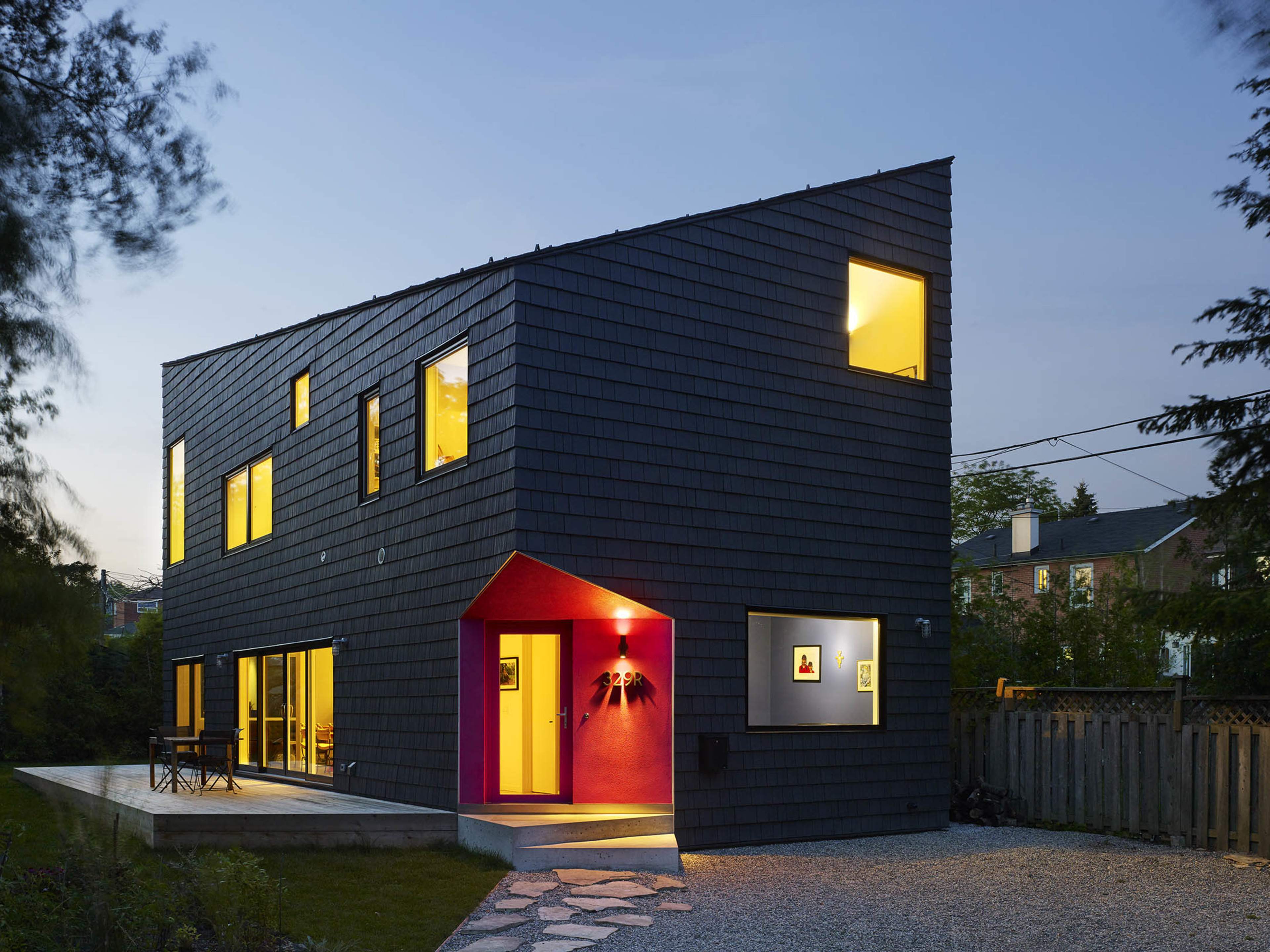 A modern two-story house with a dark shingle exterior and a prominently colored entryway, situated in a landscaped yard at dusk.