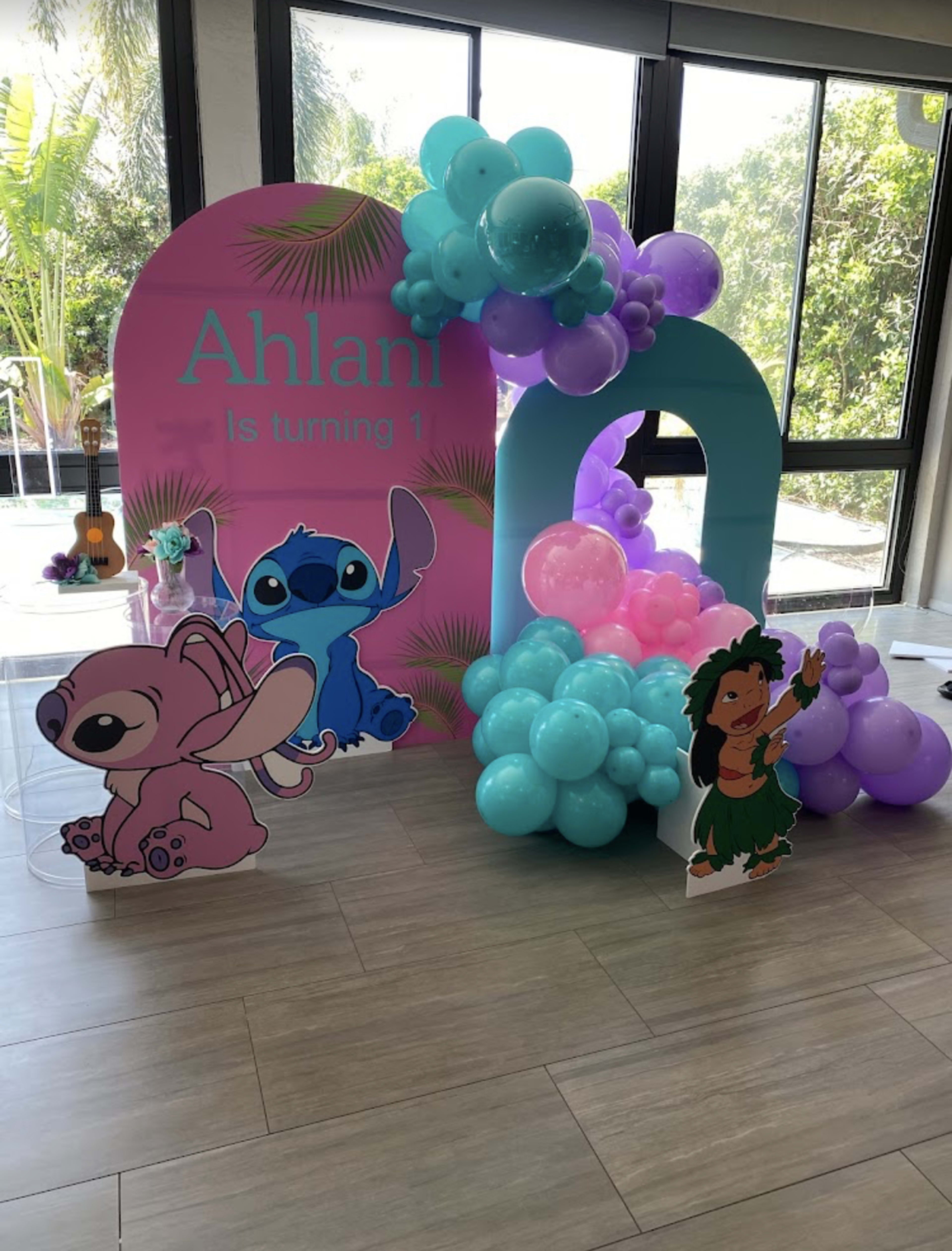 The image features a colorful birthday decoration setup for a first birthday celebration, including character displays of Stitch and Lilo, along with a balloon arch in shades of blue and pink.