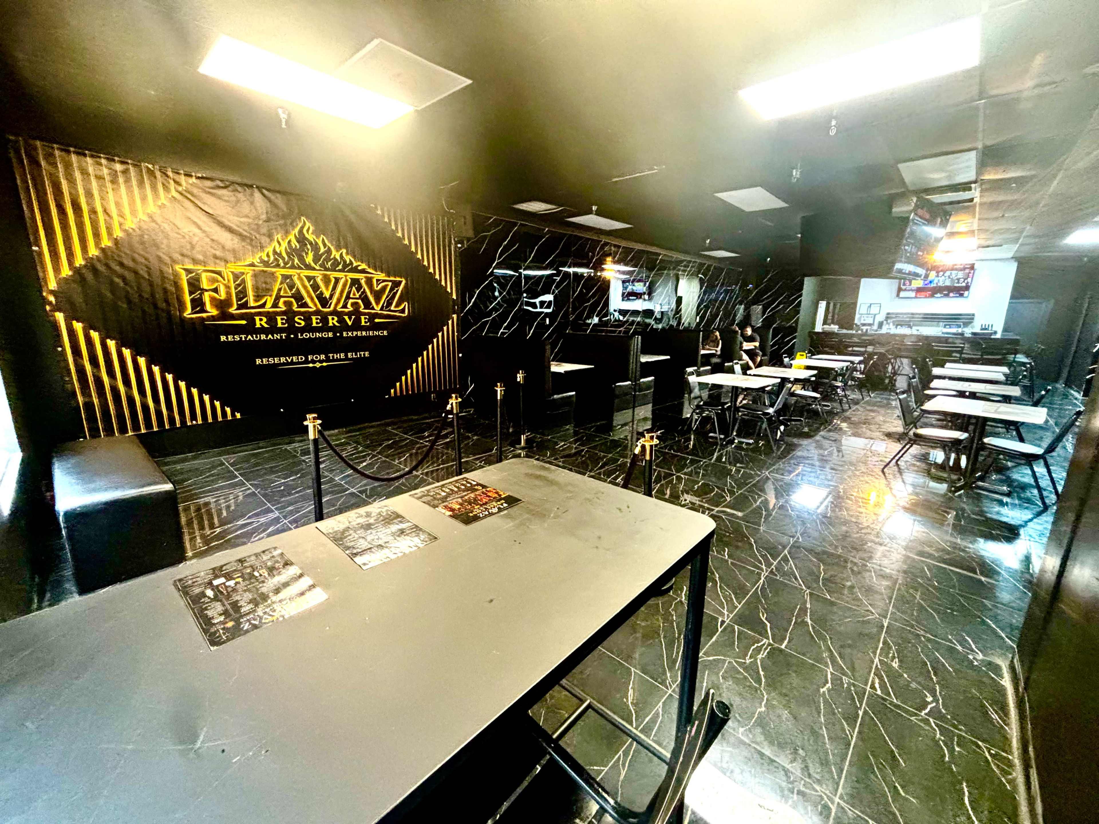 The image shows a modern restaurant interior with black marble floors, several tables, and a prominent black and gold wall featuring the name "Flavaz Reserve."