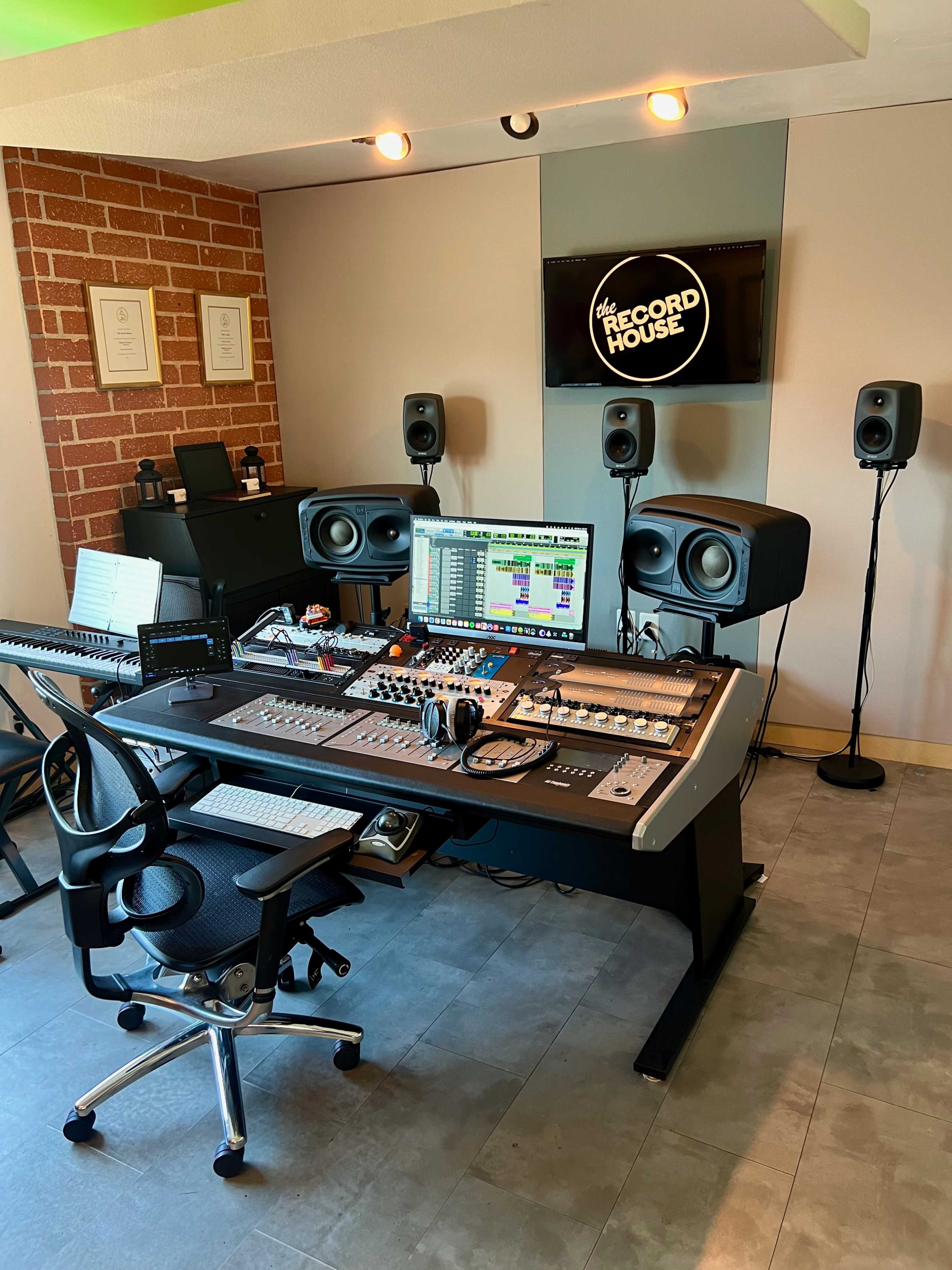 The image shows a music recording studio featuring a large mixing console, several speakers, and a computer monitor displaying audio software, all set within a modern space with exposed brick walls.