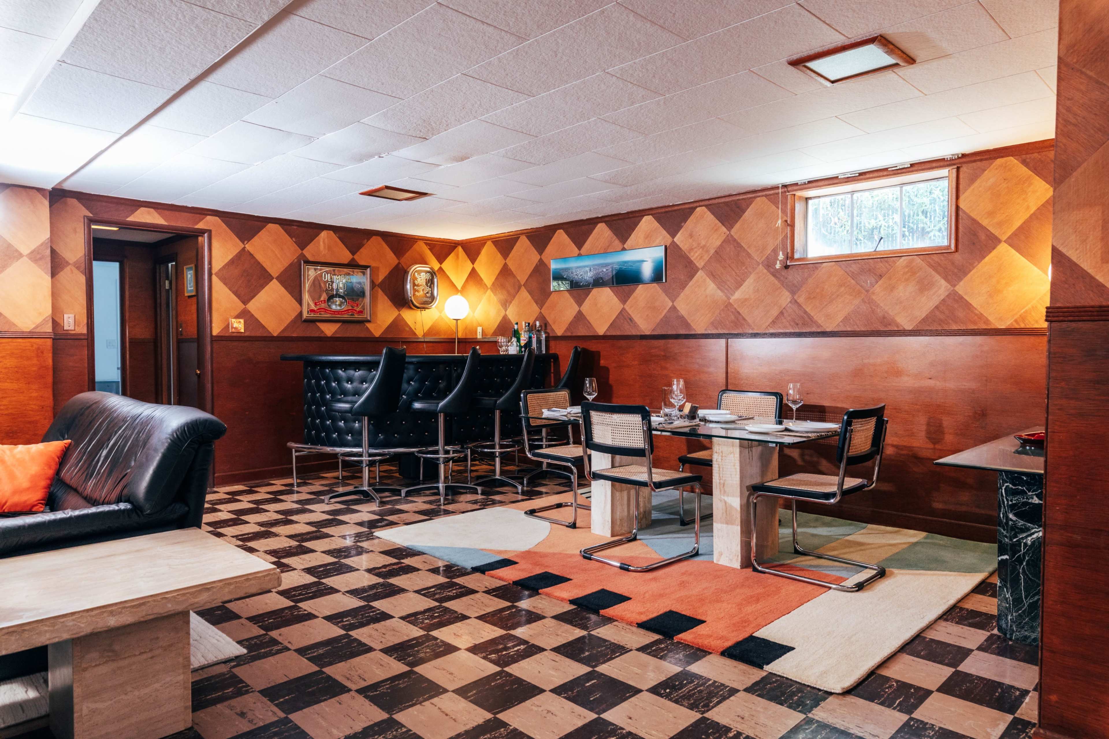 The image shows a retro-styled basement room with a black leather sofa, a dining area with a glass table and chairs, and patterned wooden walls.