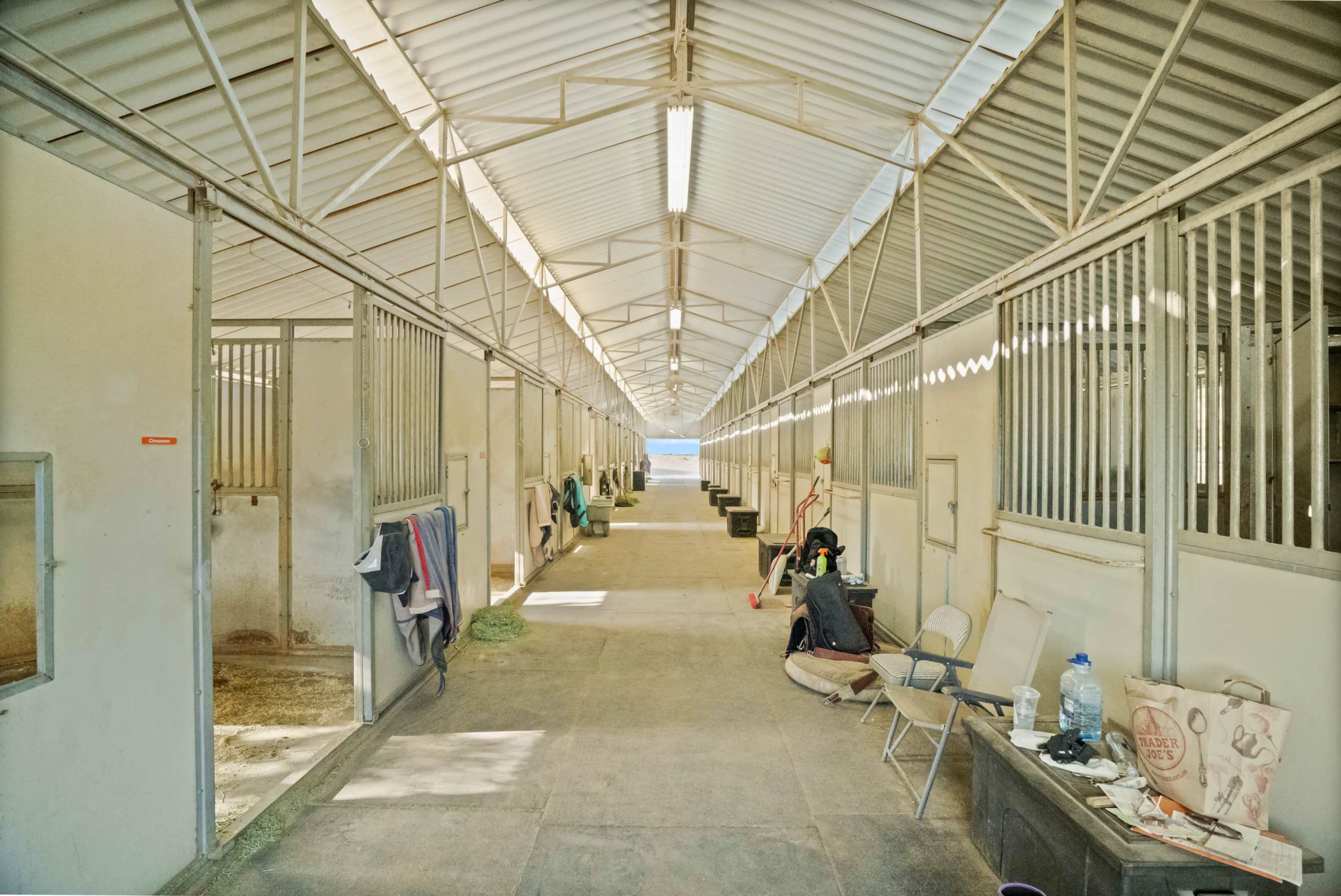 The image shows an interior view of a horse stable with empty stalls arranged along a long corridor.