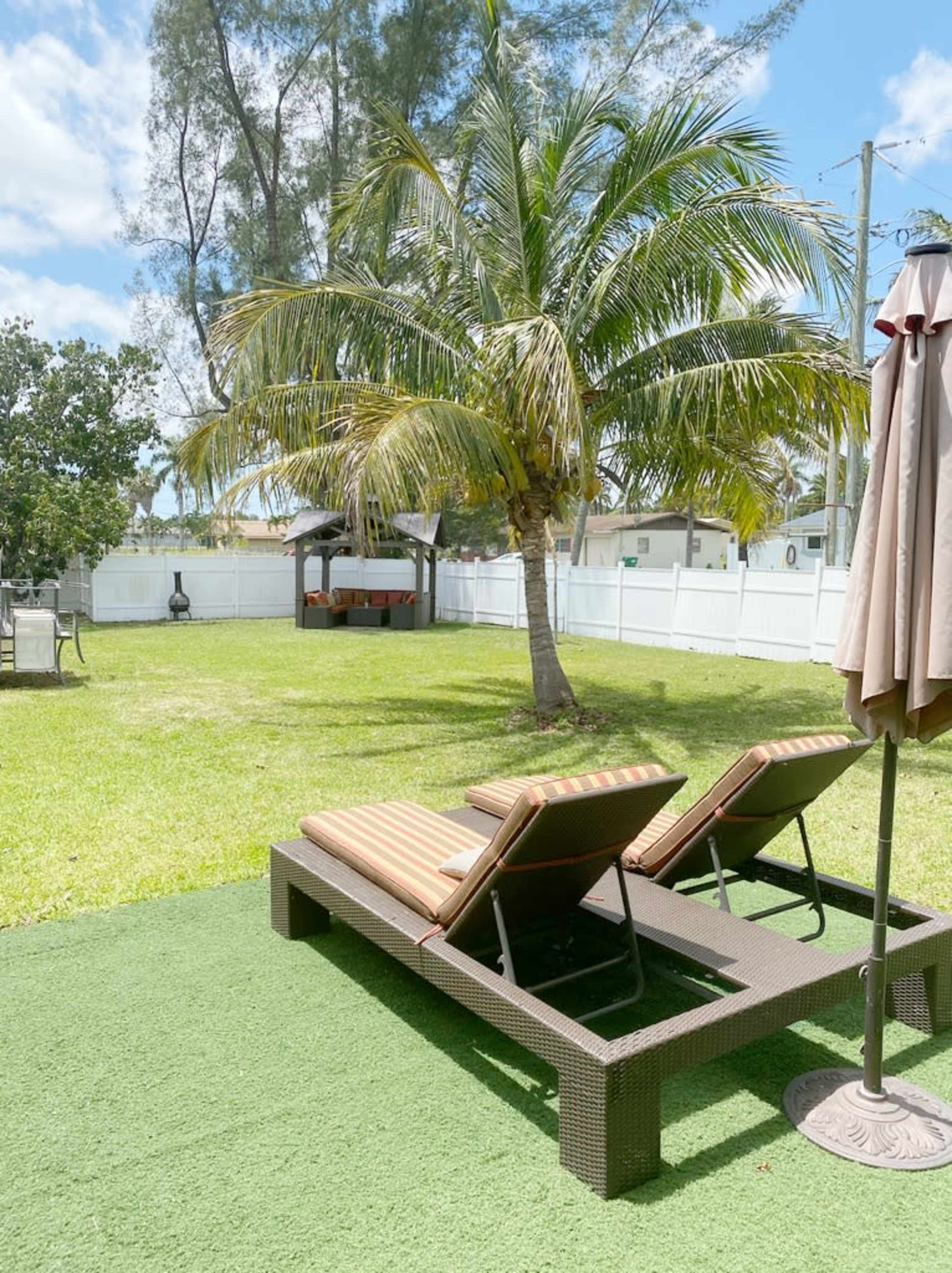 A backyard with two lounge chairs under a palm tree, surrounded by green grass and a white fence.