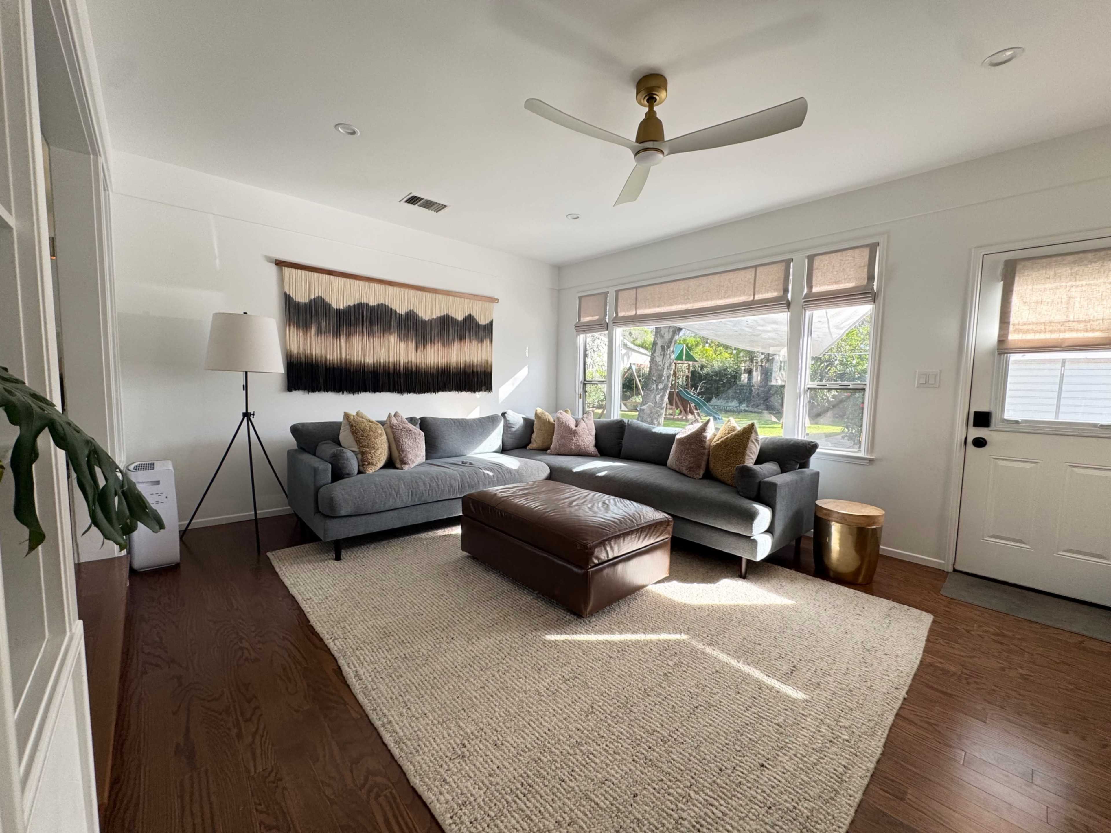 The living room features a gray sectional sofa with decorative pillows, a brown leather ottoman, a light-colored area rug, and large windows allowing natural light to fill the space.