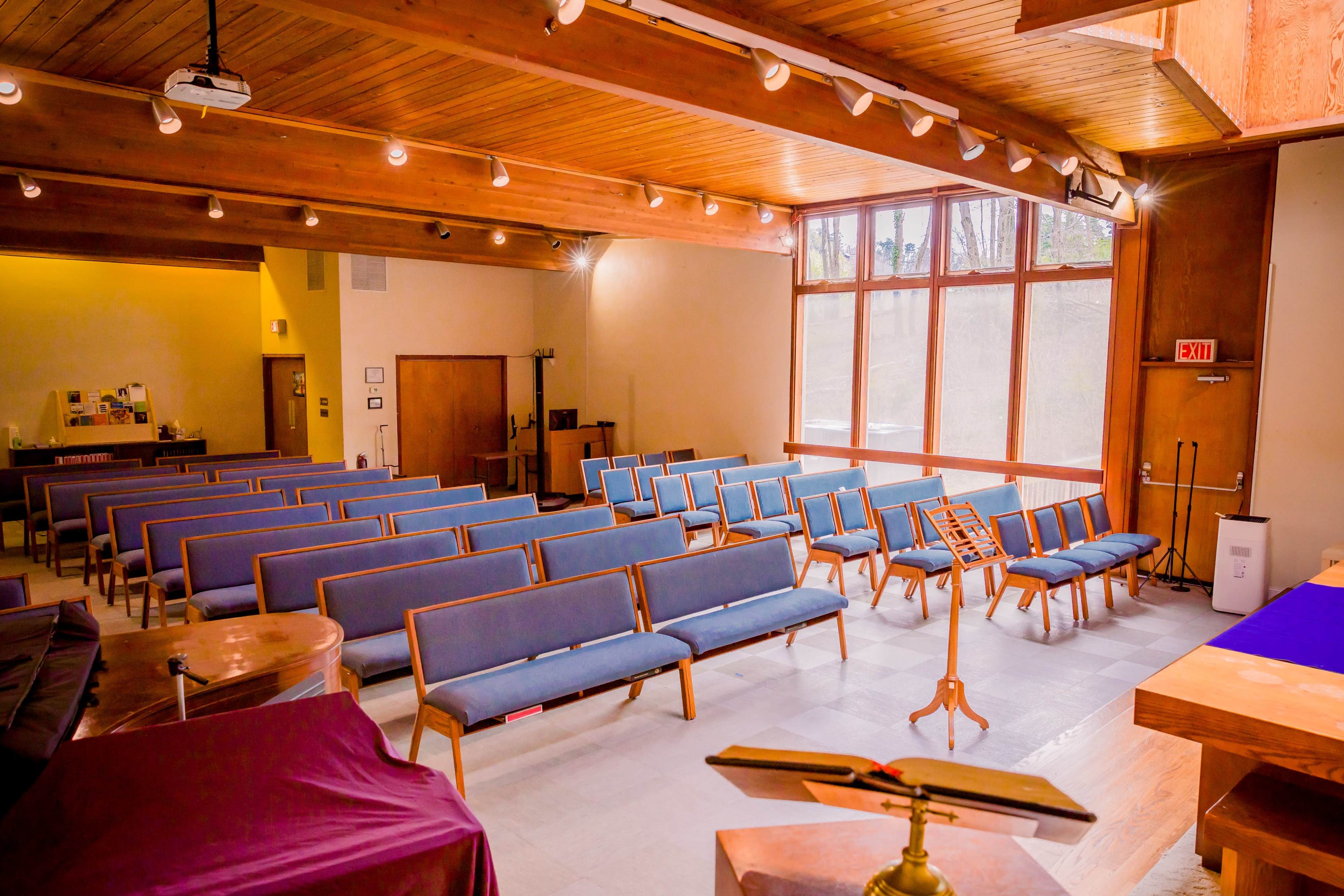 The interior of a chapel features rows of blue pews, large windows letting in natural light, and a pulpit with a music stand.