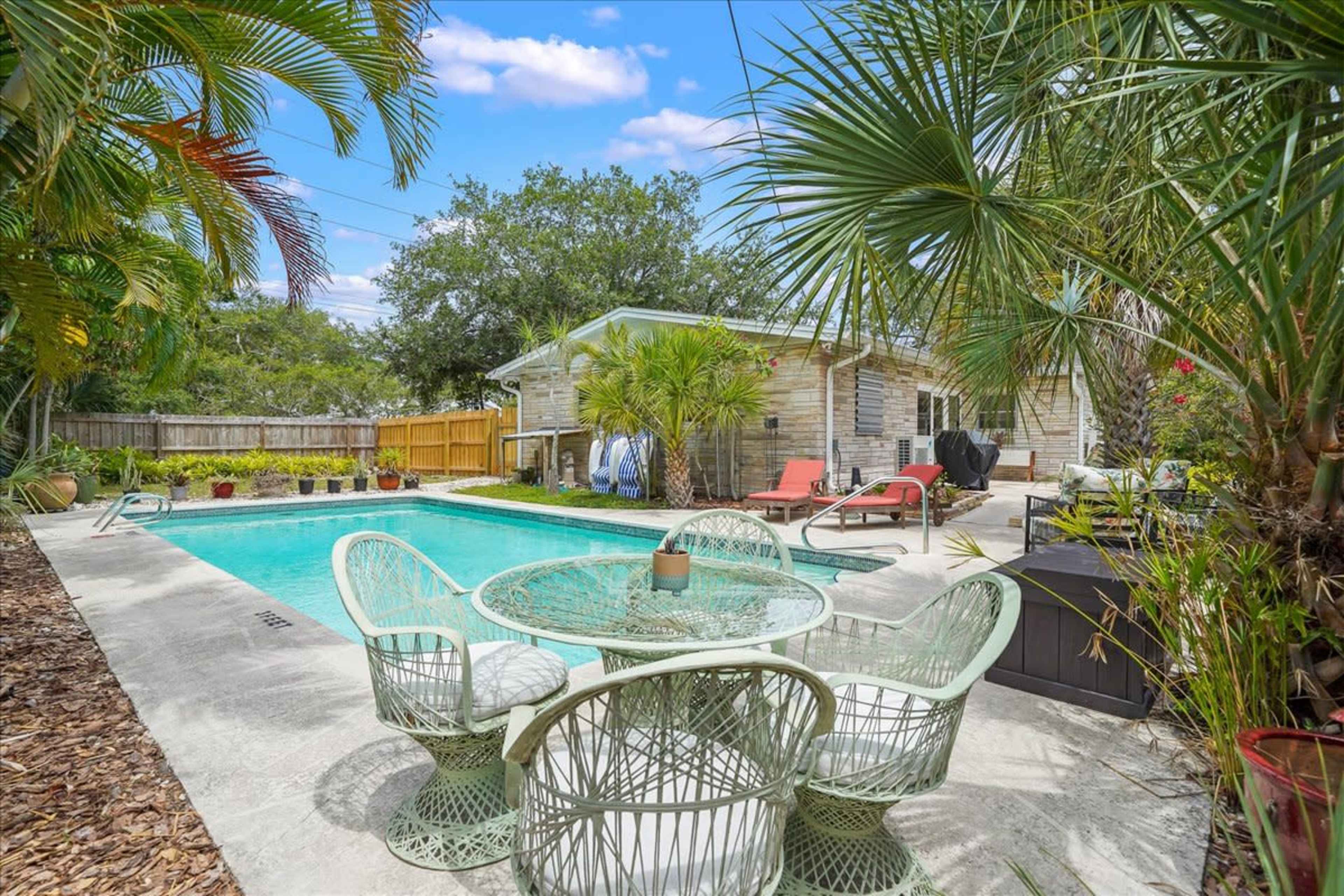A backyard with a swimming pool surrounded by palm trees and a seating area featuring a glass-top table and four chairs.