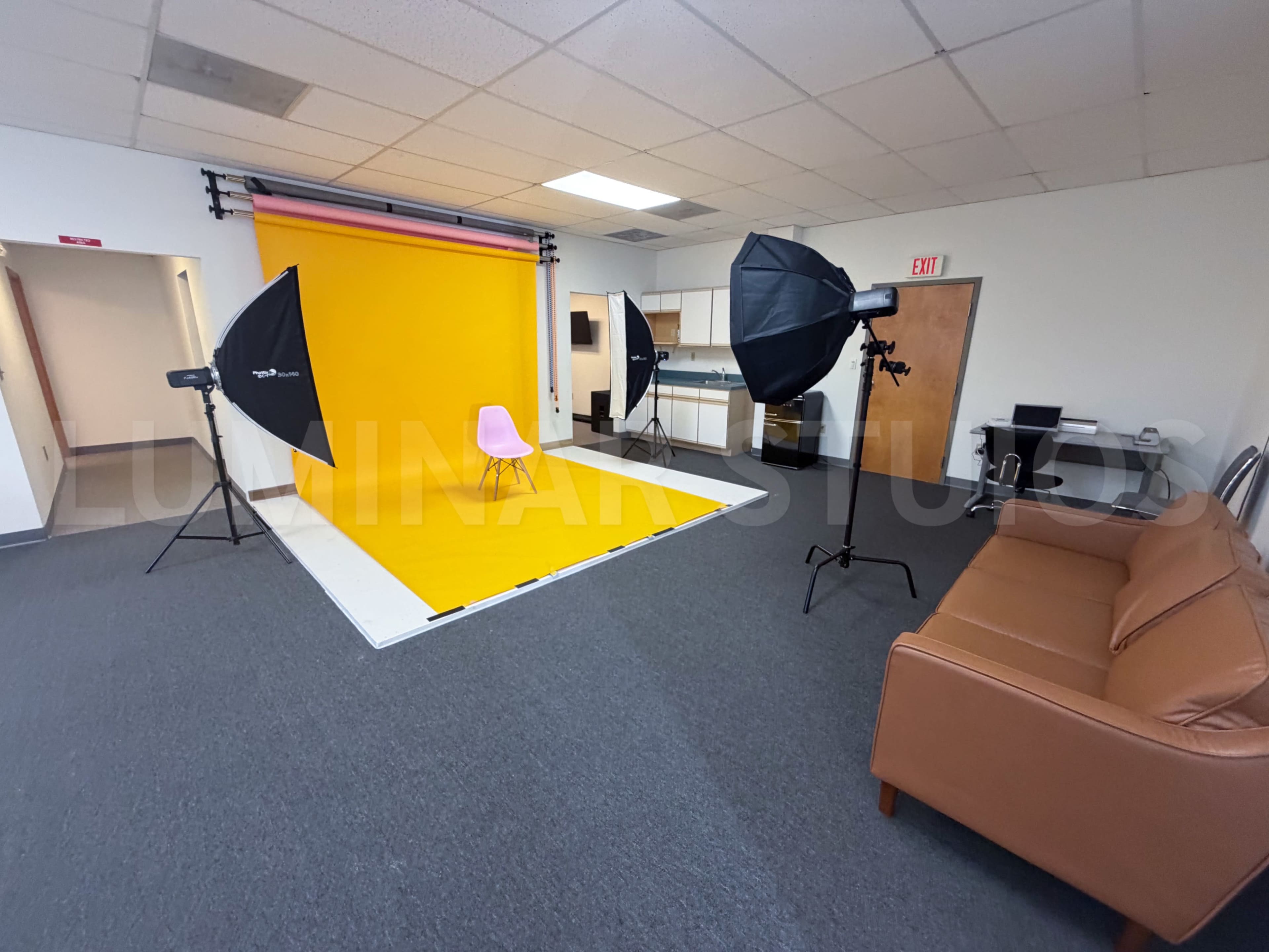 The image shows a photography studio with a bright yellow backdrop, softbox lighting, a pink chair, and a brown couch in the corner.