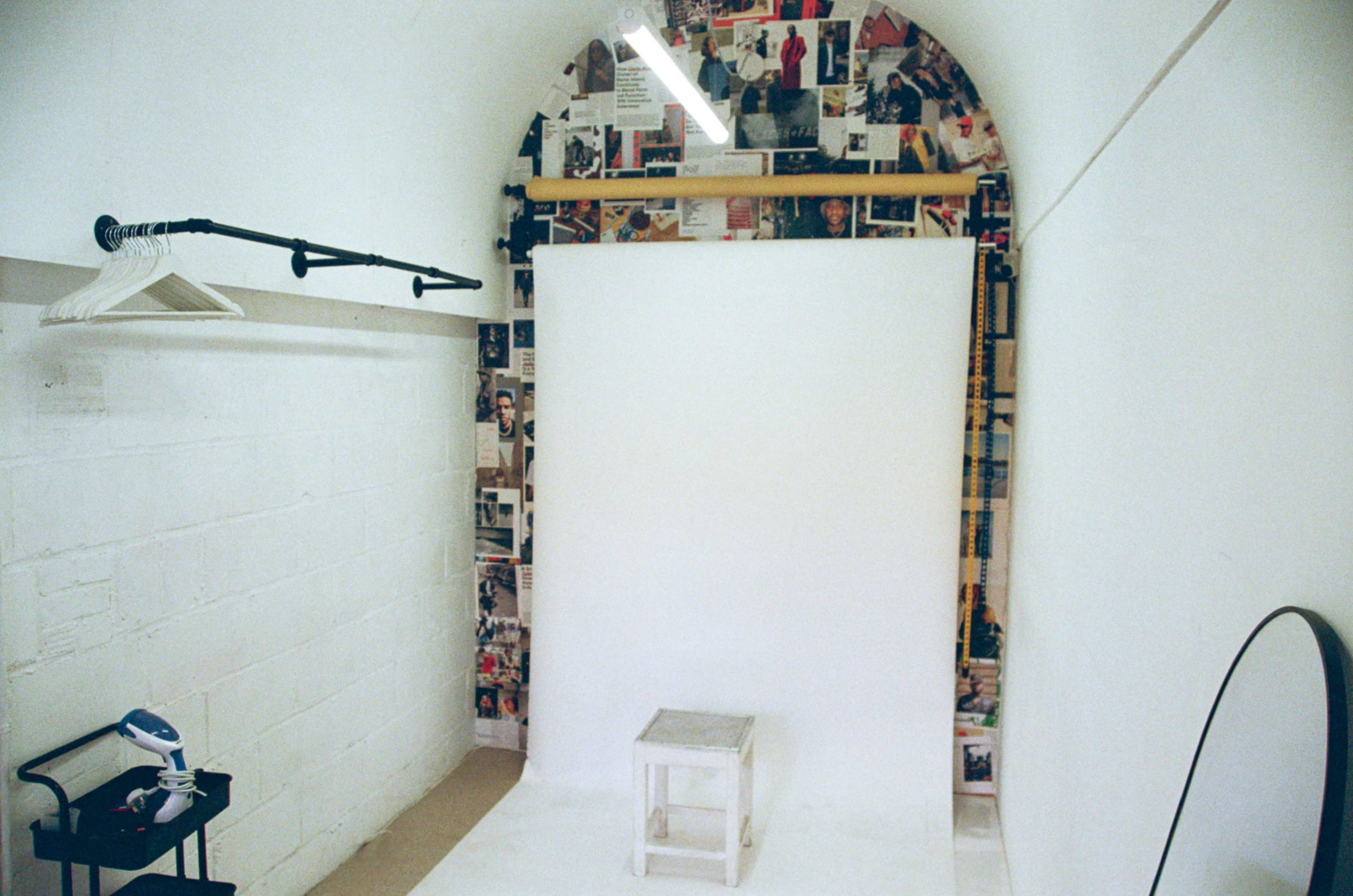 The image shows a small photography studio with a white backdrop, a stool, a hanging clothes rack, and a wall covered in various magazine clippings.