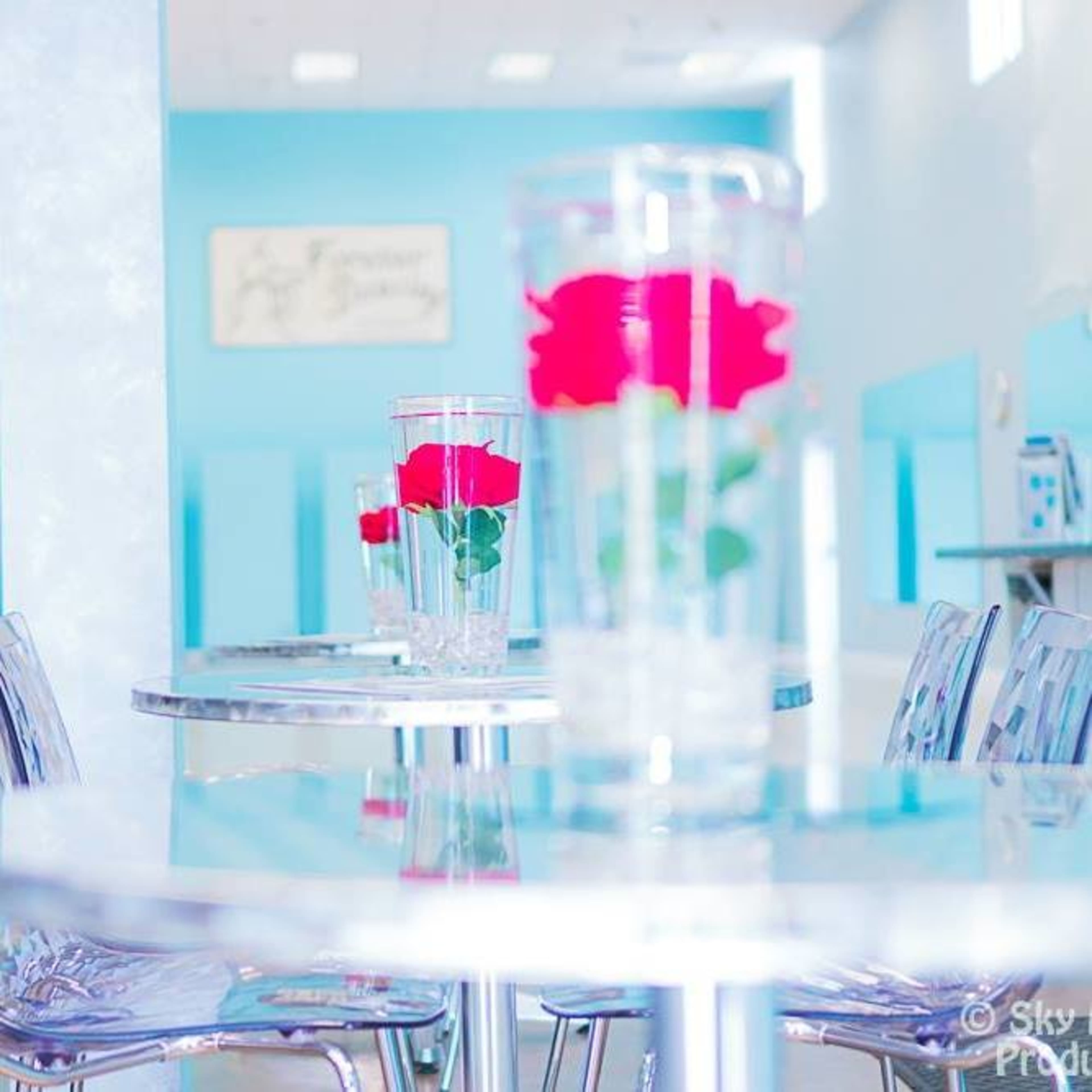 Transparent tables with glasses containing roses, set against a light blue wall in a bright, modern space.
