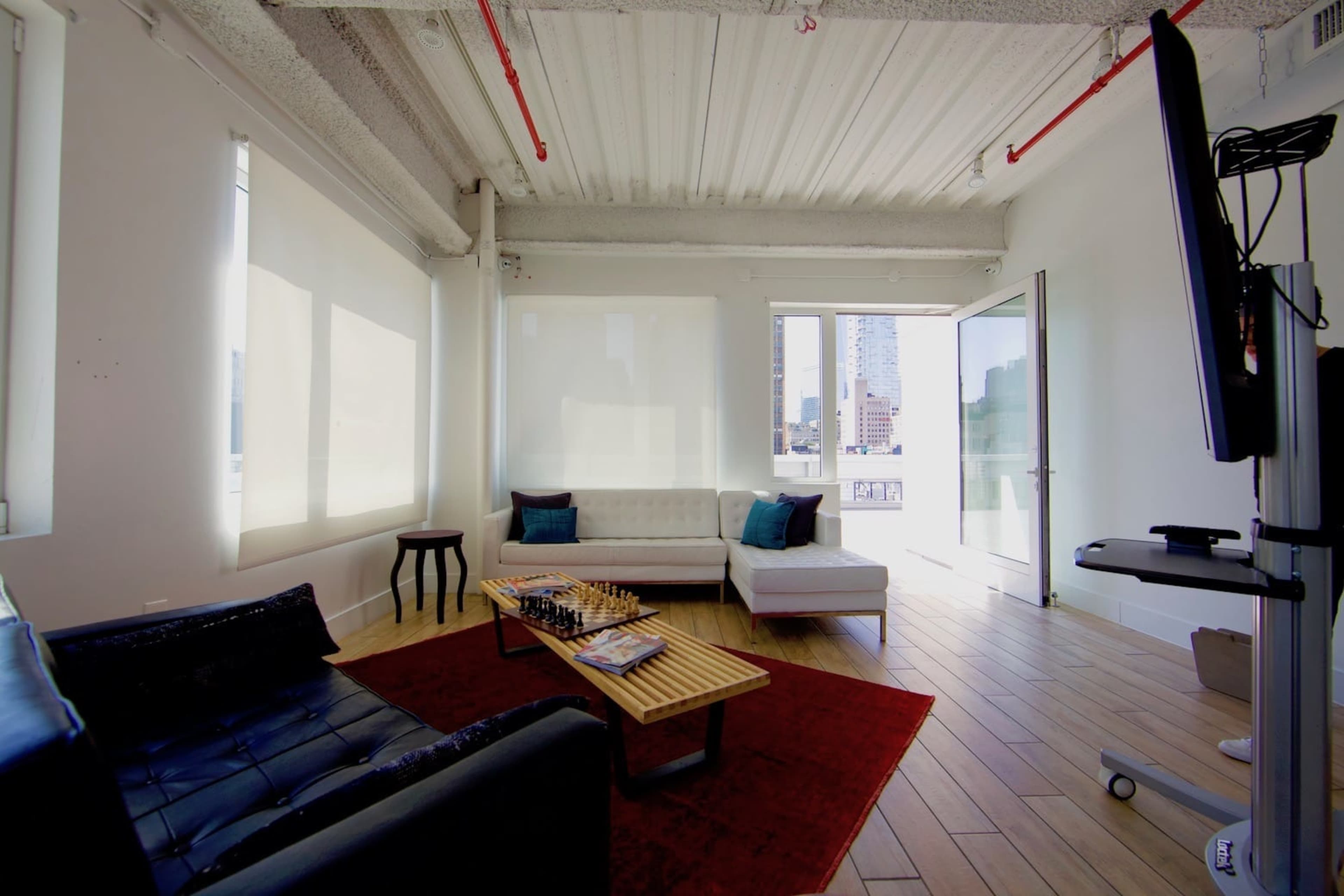 A modern office space with a seating area that includes a black sofa, white sectional, a chessboard on a wooden coffee table, and large windows overlooking the city.