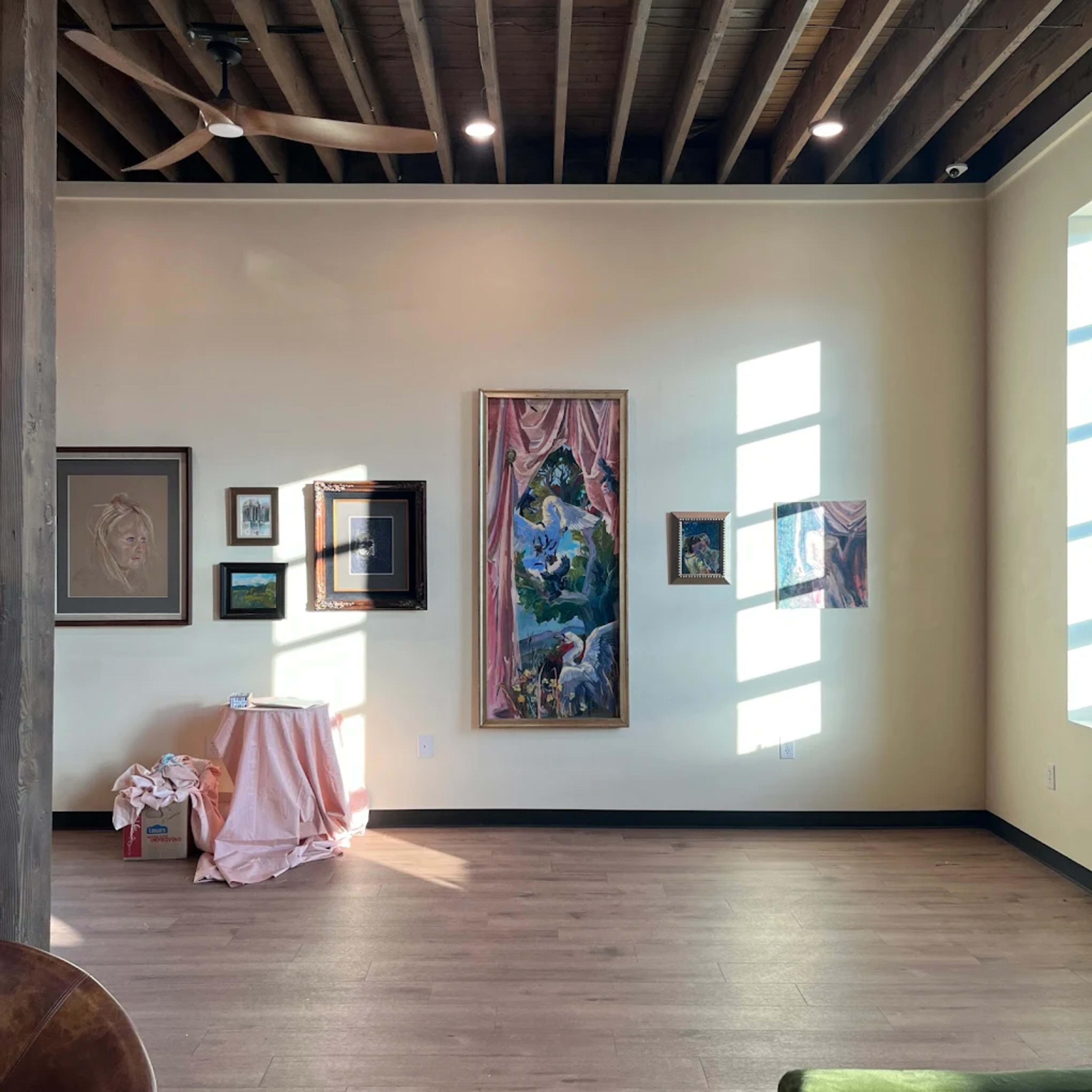 The image shows a well-lit interior space featuring a collection of framed artworks on the walls, with a small table covered in fabric and a box in the corner.