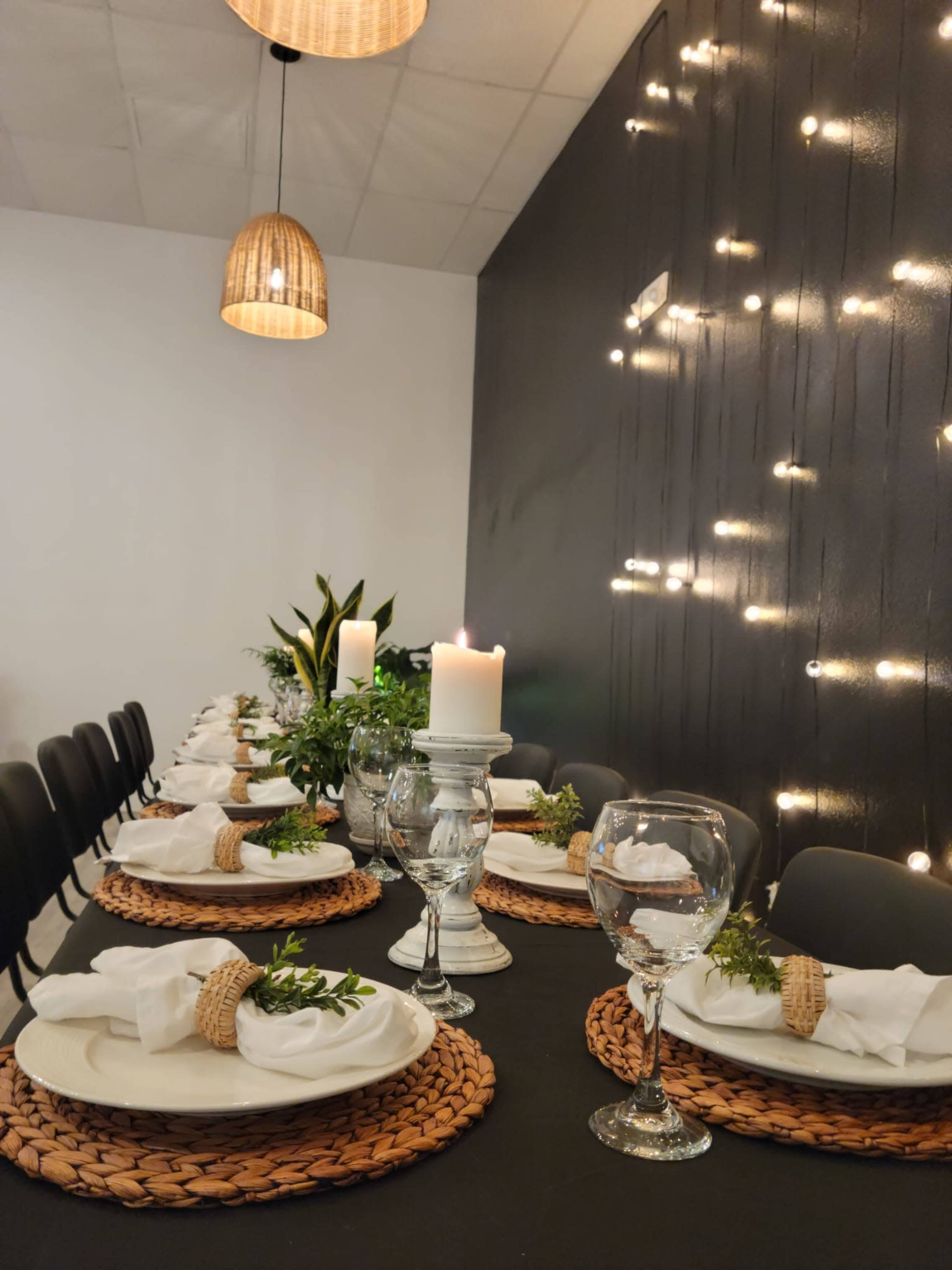 A long dining table is set with white plates, woven placemats, glasses of water, and candles, illuminated by pendant lights and string lights on a dark wall.