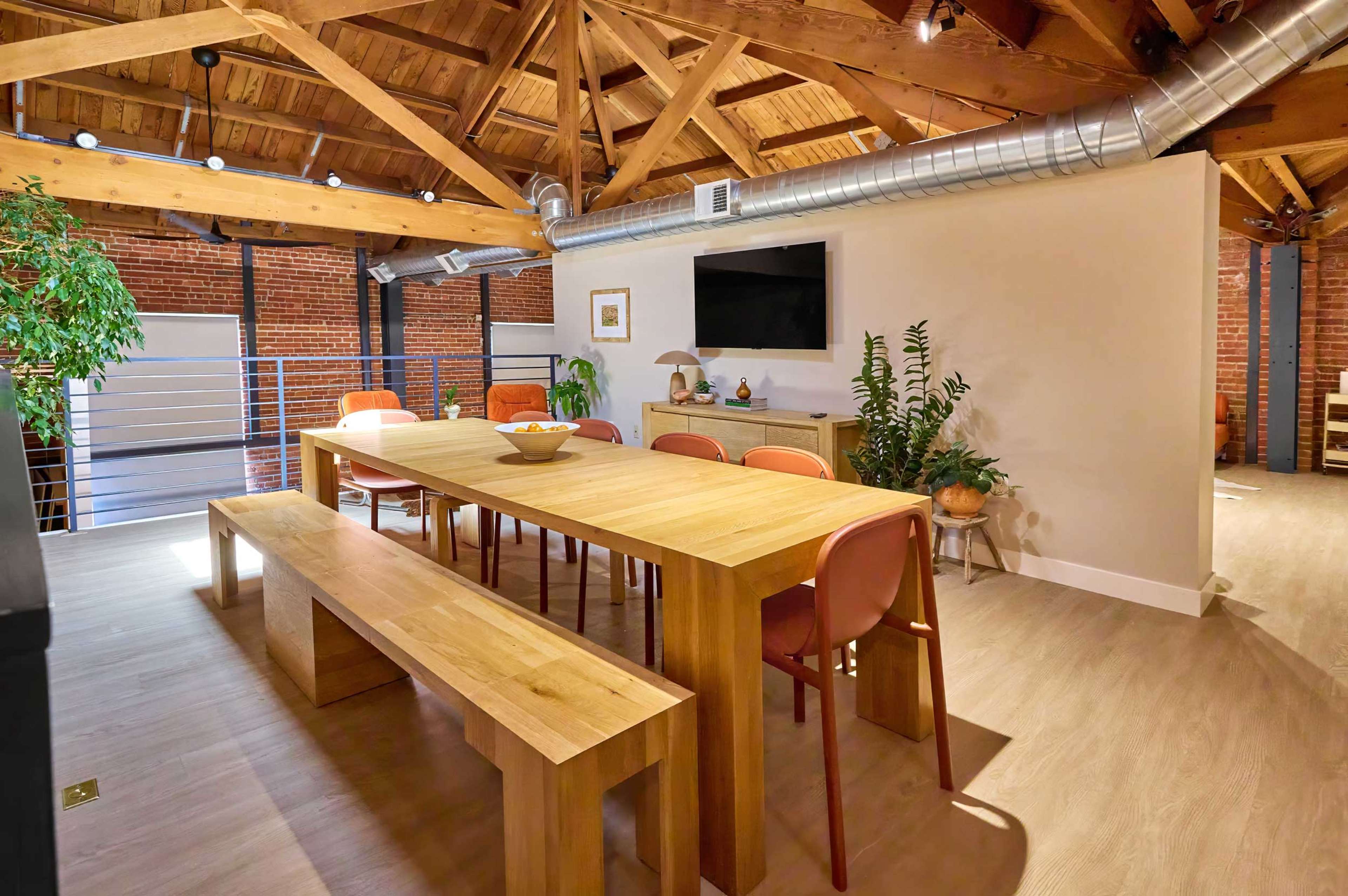 The image shows a dining area with a large wooden table and bench, surrounded by orange chairs, set in a room that features exposed wooden beams and brick walls.
