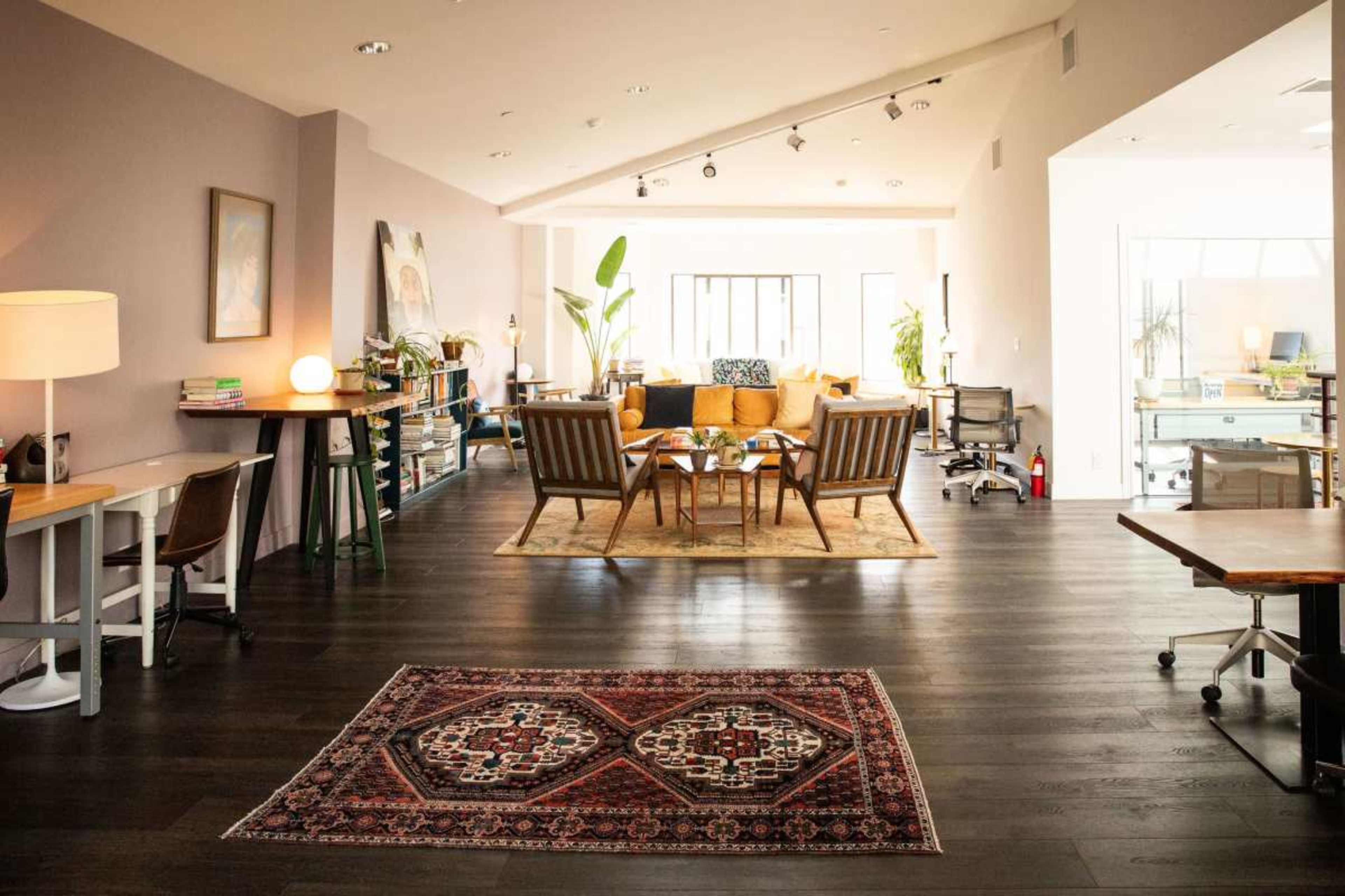 The image shows a modern open-plan office space featuring a seating area with a large rug, a coffee table, and various desks arranged throughout the room.