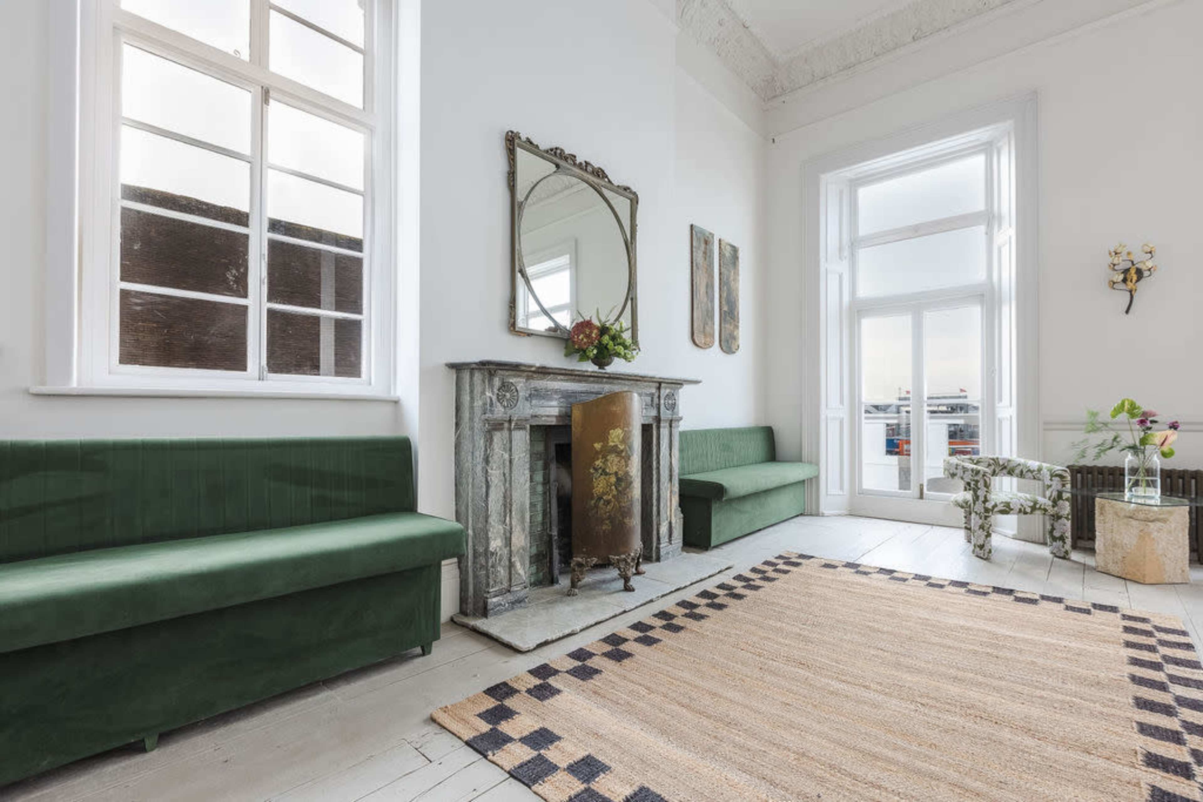 A bright room featuring green velvet benches, a marble fireplace, a large mirror, and a patterned area rug.