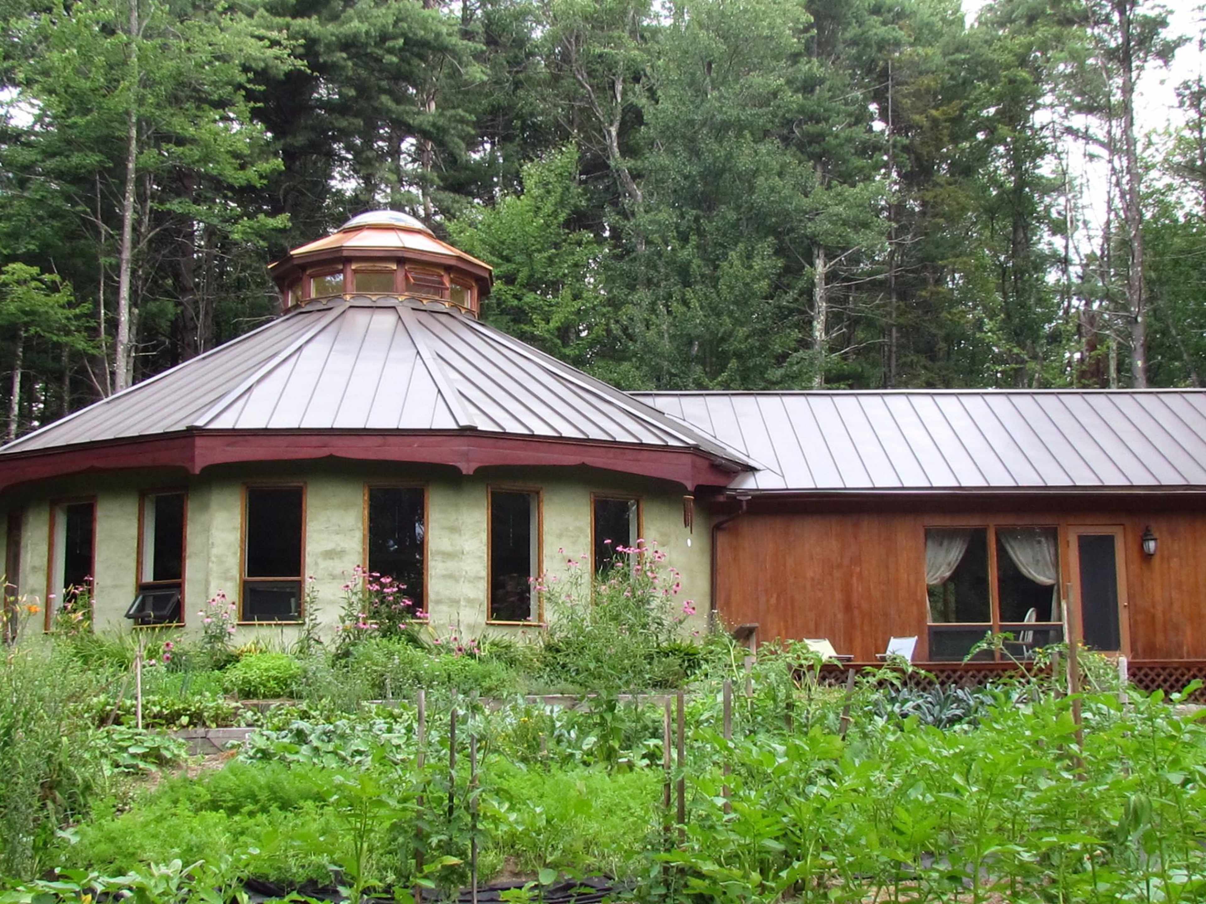 A round house with a copper dome and large windows is connected to a wooden structure and surrounded by a vegetable garden in a forested area.