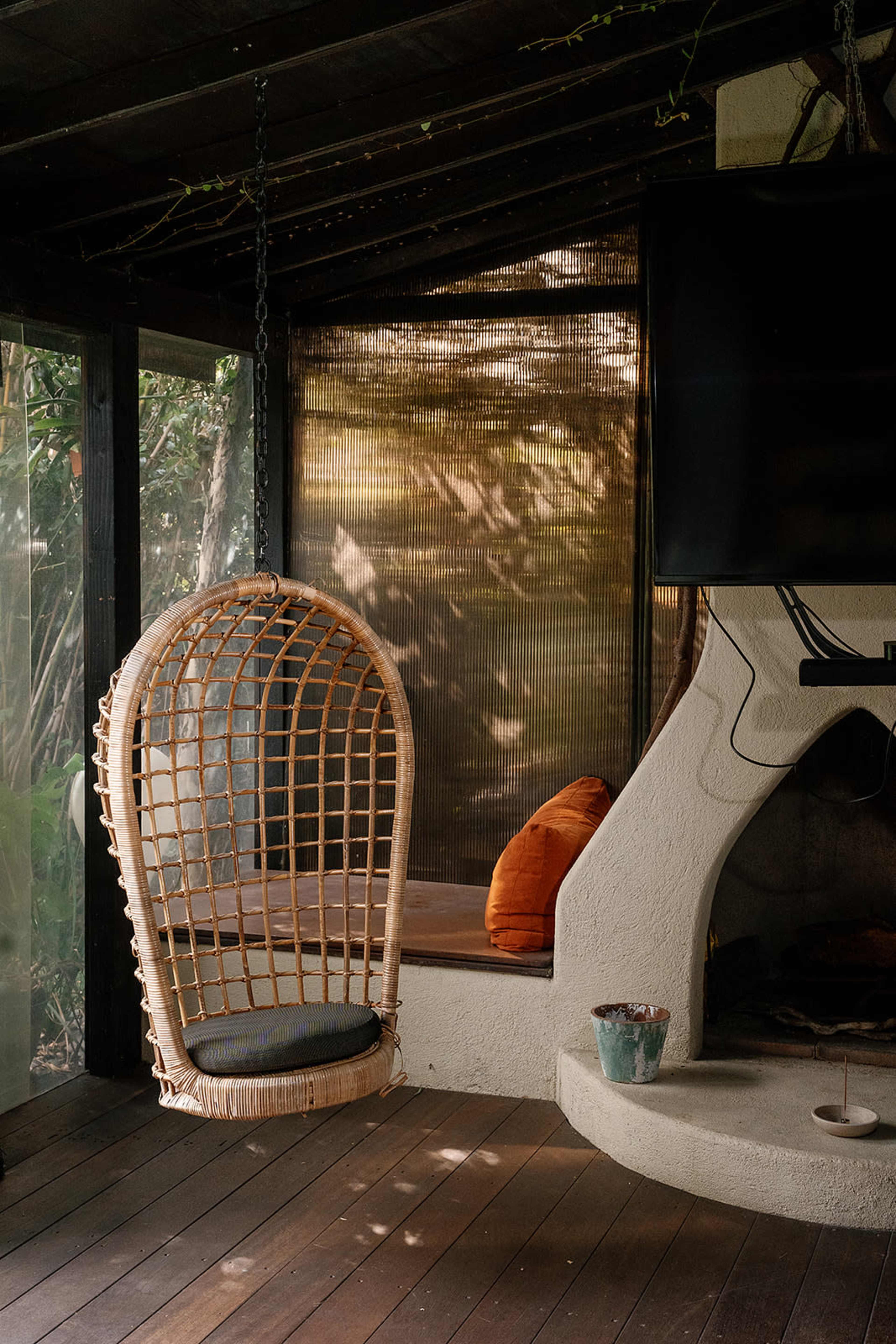 The image depicts a cozy interior with a hanging rattan chair, a modern fireplace, and large windows overlooking a green outdoor space.