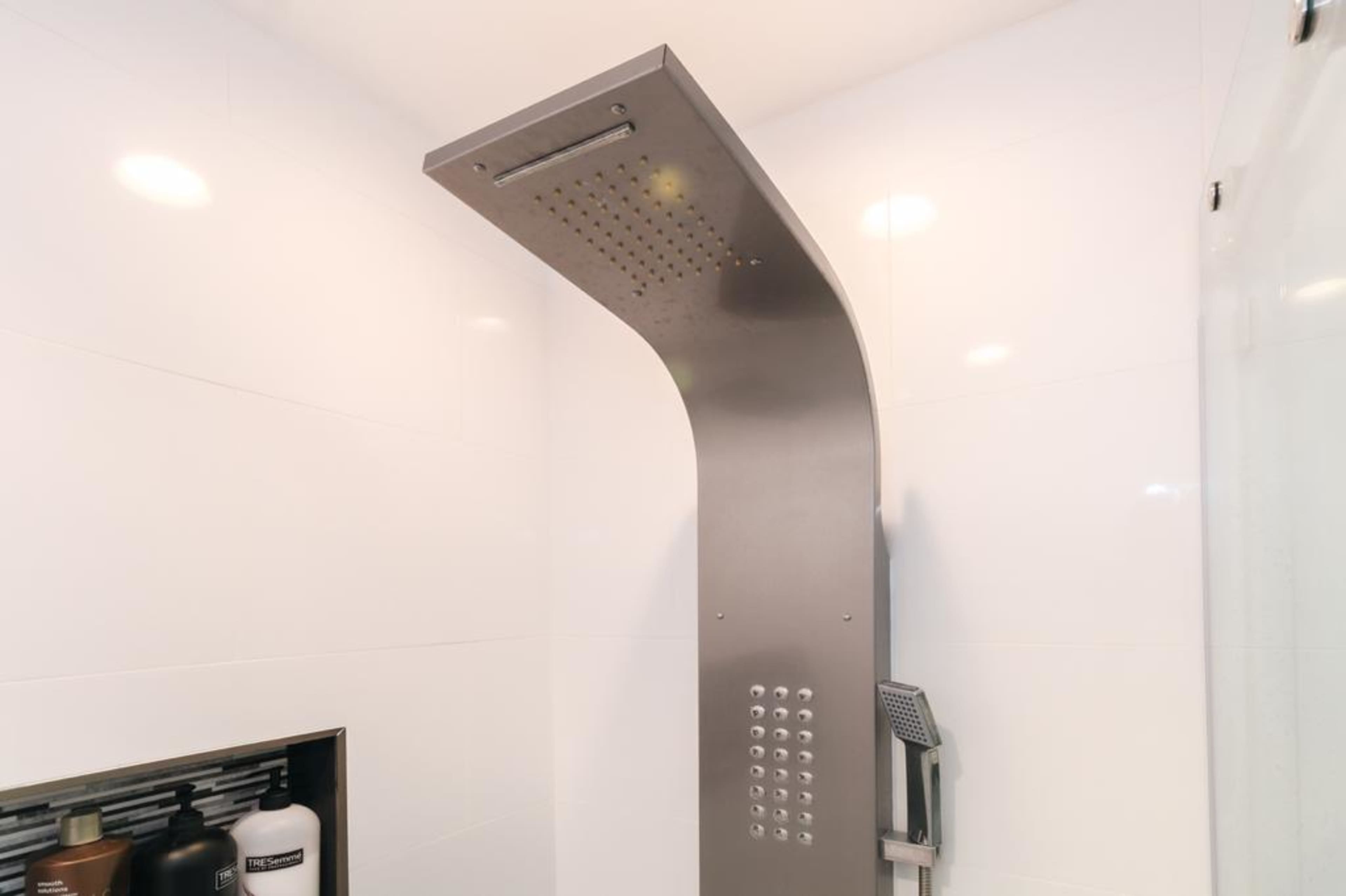 The image shows a modern shower structure with a sleek, curved metallic showerhead mounted on a wall, alongside a shelf containing toiletries.