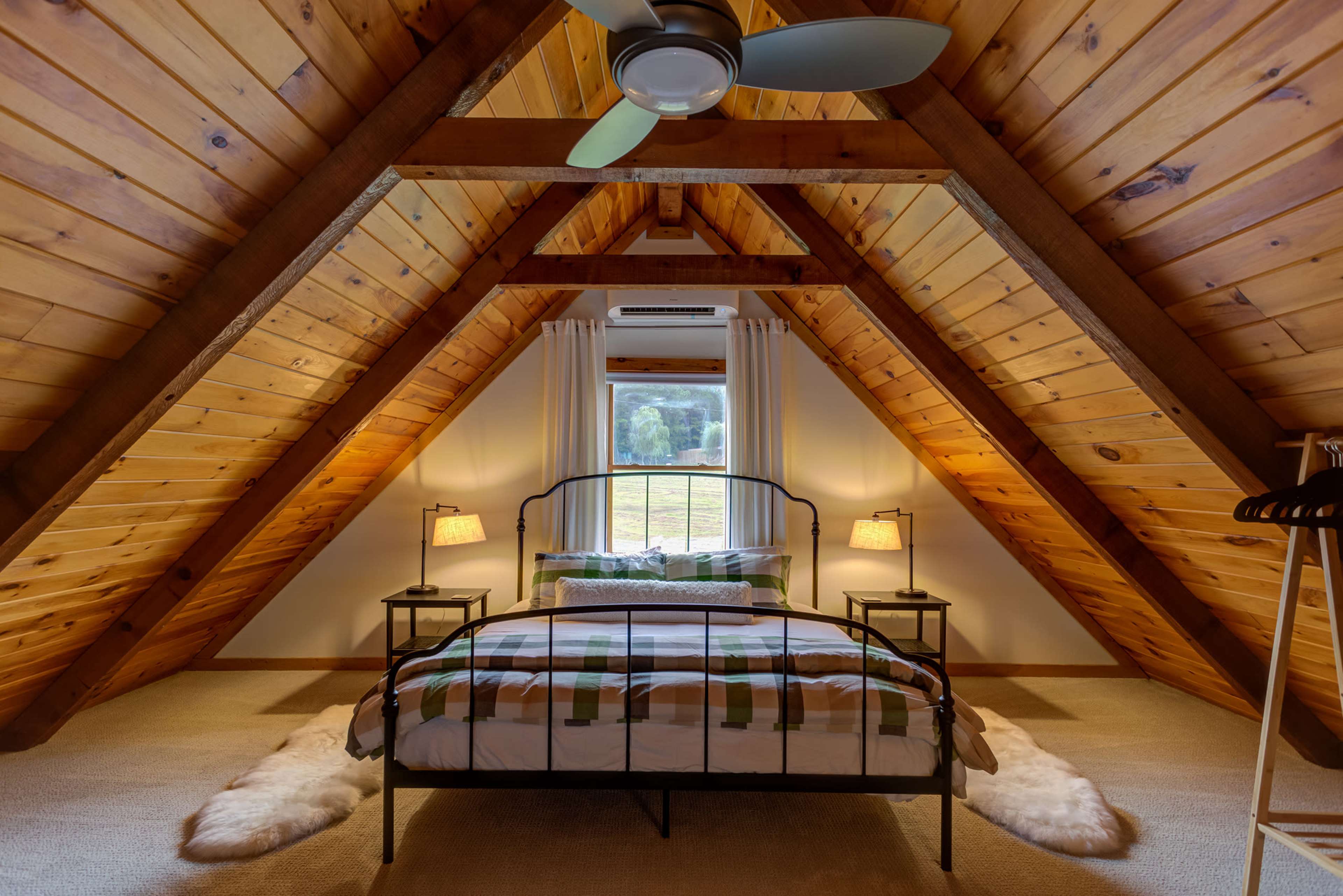A cozy attic bedroom featuring a metal bed frame with a patterned comforter, two bedside lamps, and a large window with a view of the outdoors, all under a wooden A-frame ceiling.